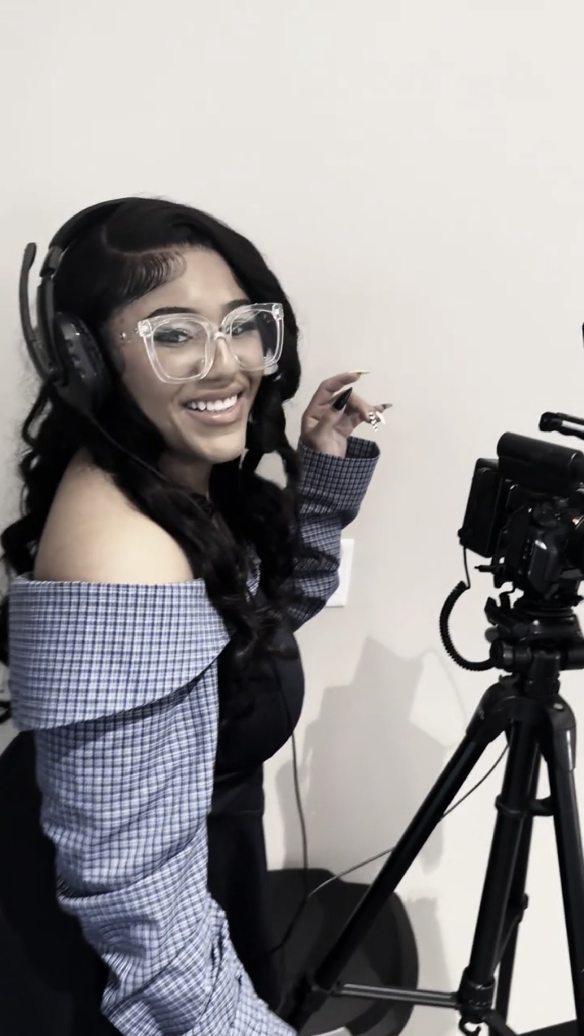 Woman with glasses and headphones smiling at camera, standing next to tripod with camera, against plain white wall.