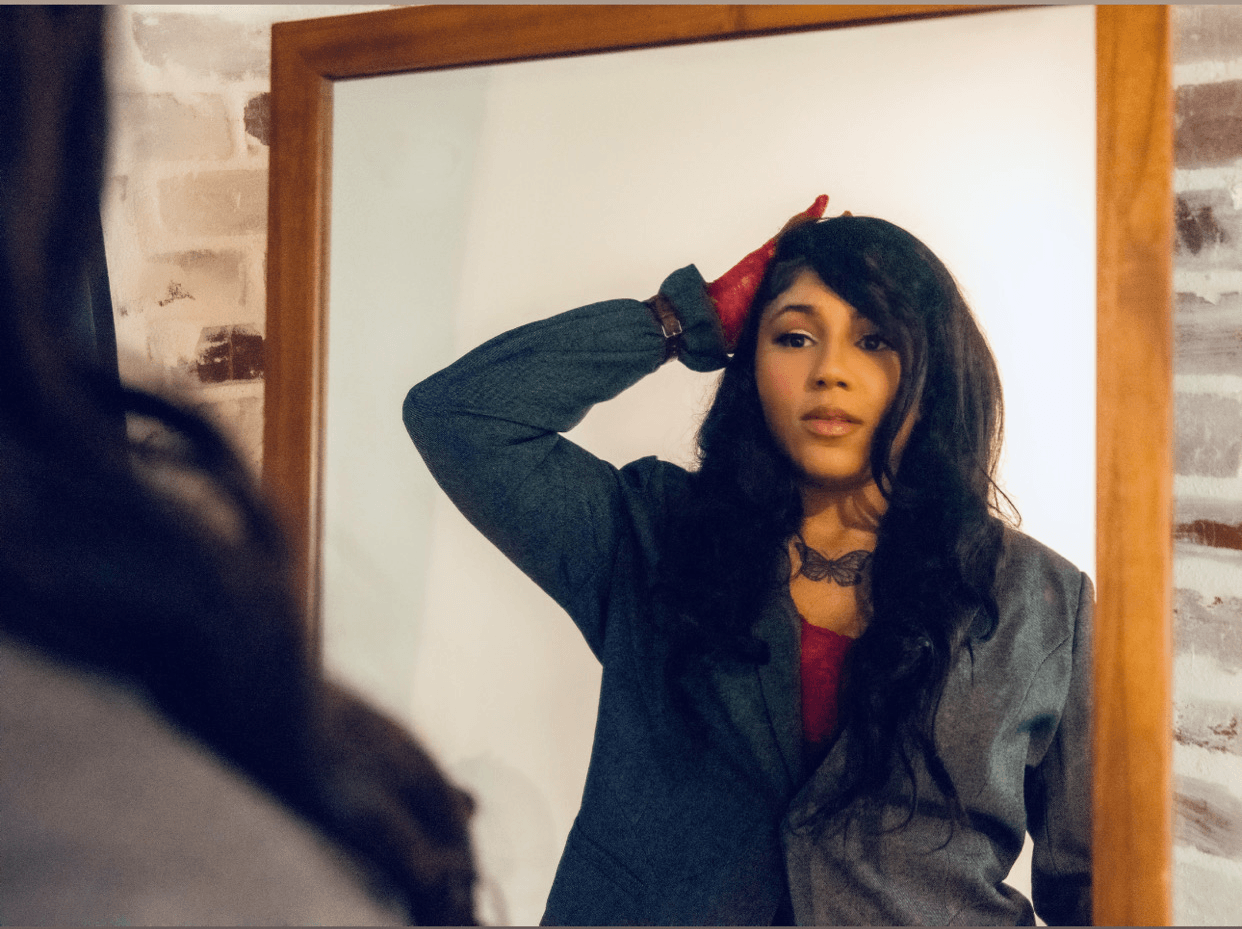 Woman with long dark hair, wearing a blazer, standing in front of a whiteboard, with a brick wall background.
