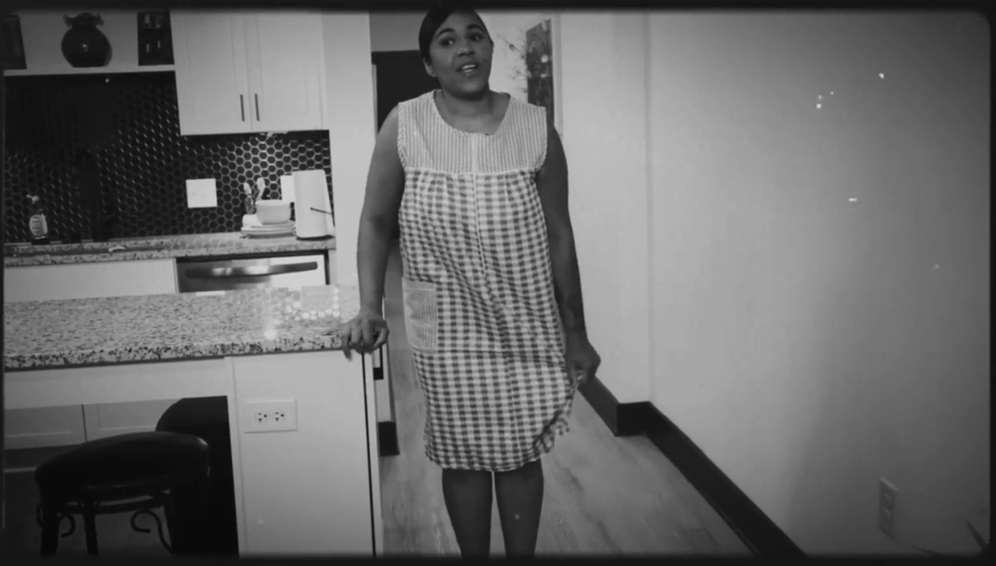 Woman standing in a kitchen, wearing a checkered apron over a dress, with cabinets and a countertop behind her.