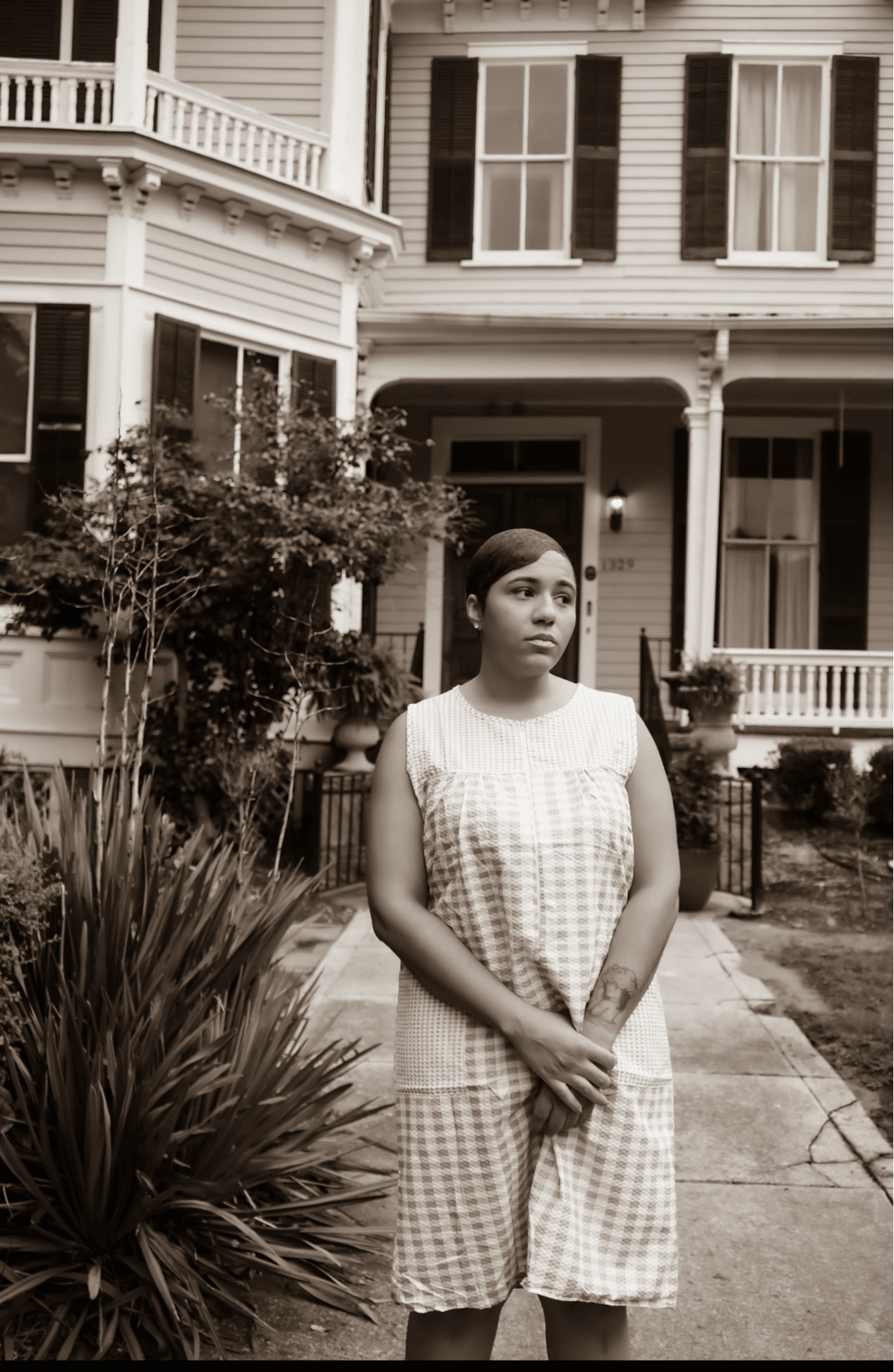 Young woman stands in front of a house with a porch, garden, and plants, looking to the side.