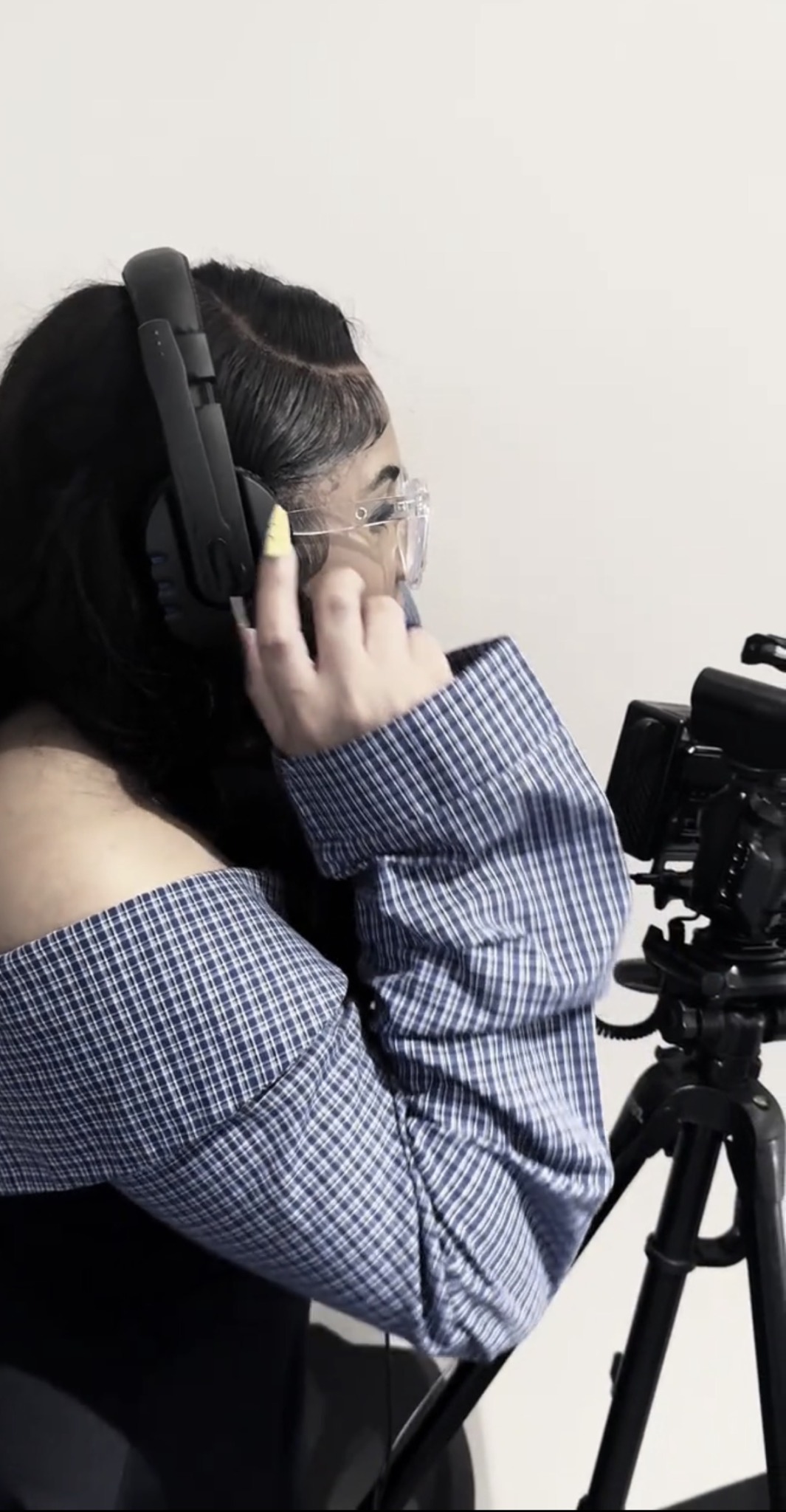Person wearing headphones and glasses, adjusting equipment on a tripod, facing right, against a plain background.