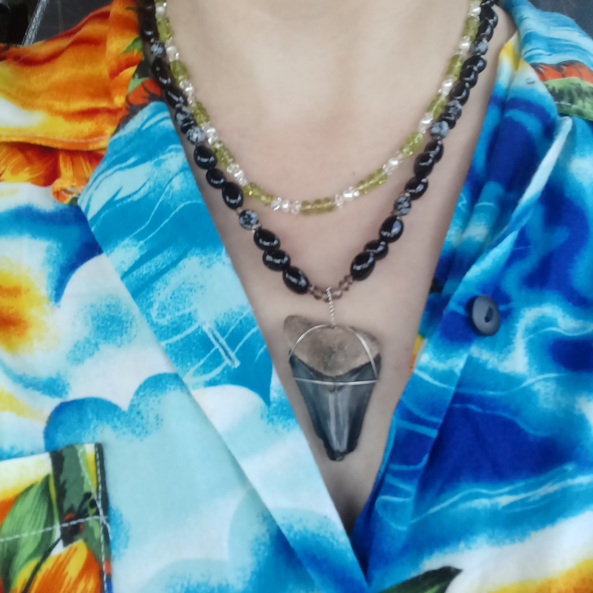 Person wearing beaded necklace with a large, pointed, gray and black pendant, dressed in colorful, patterned clothing.