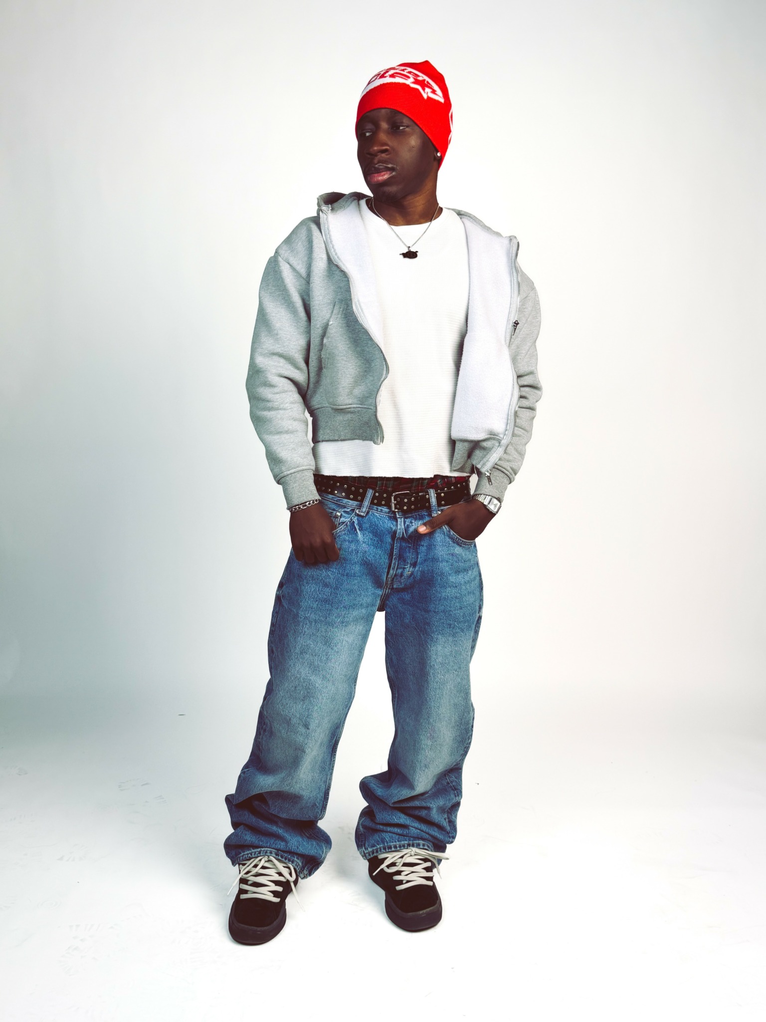 Young man wearing a red beanie, gray hoodie, white shirt, and jeans, standing with hands in pockets against a plain background.