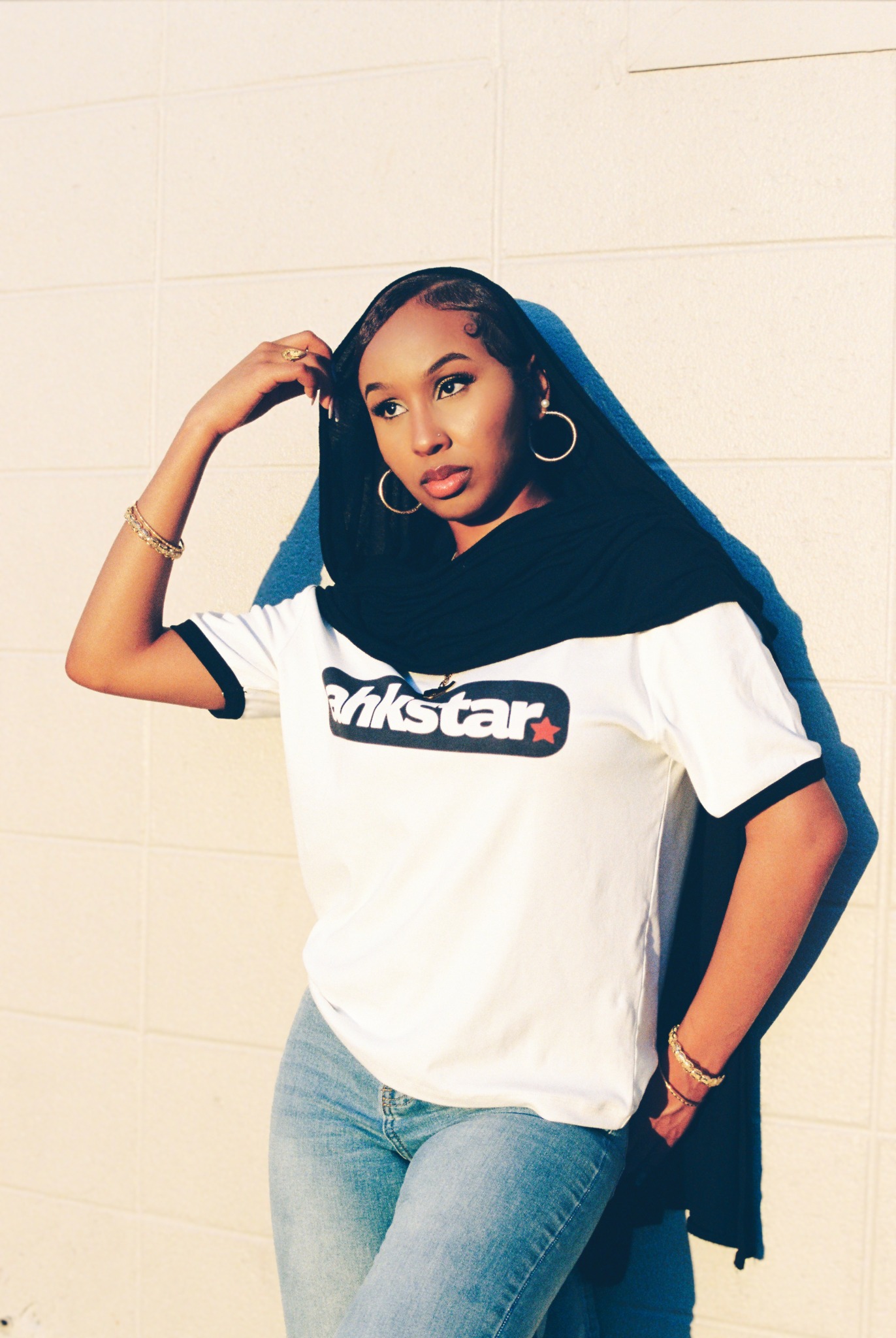 Young woman with long hair, wearing a white and black t-shirt, jeans, and hoop earrings, standing against a white wall.