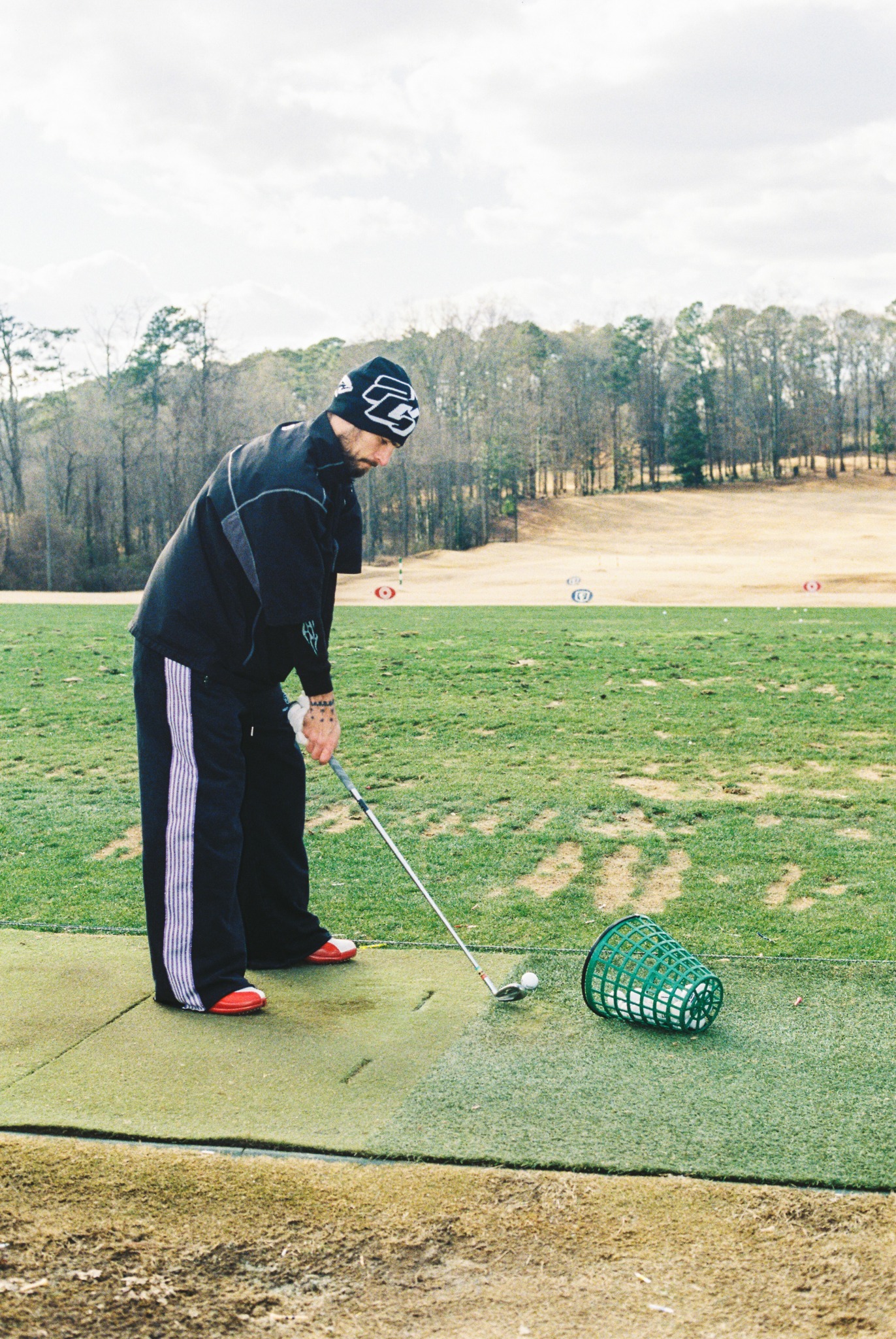 Person preparing to hit golf ball on tee box with basket nearby, grassy field and trees in background.