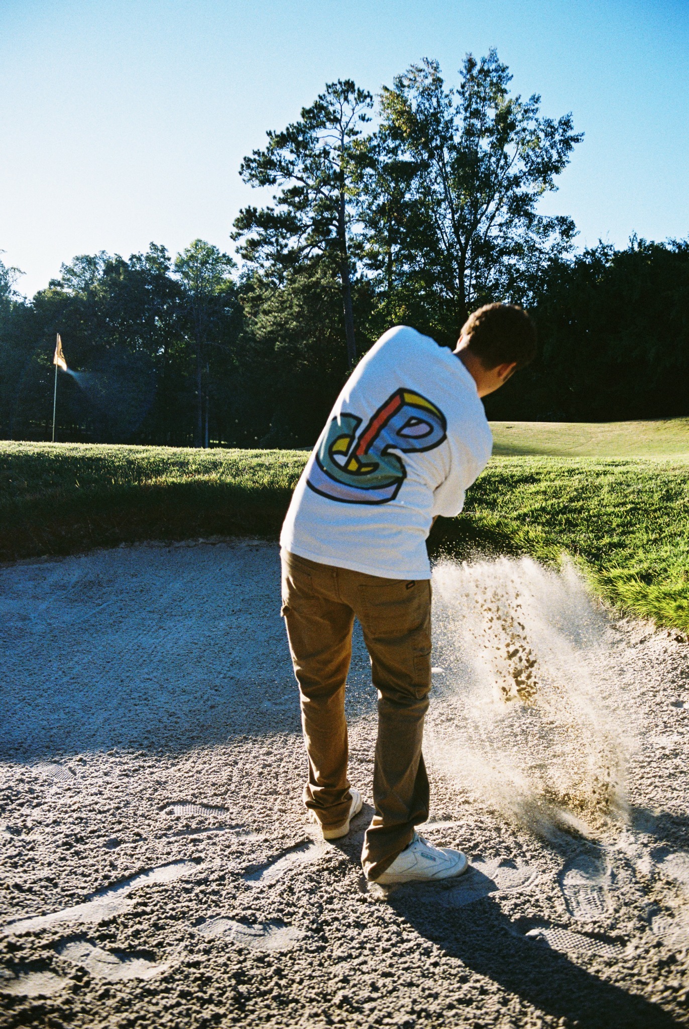 Person in white shirt and brown pants standing in a sand bunker on a golf course, with trees and a flag in background.
