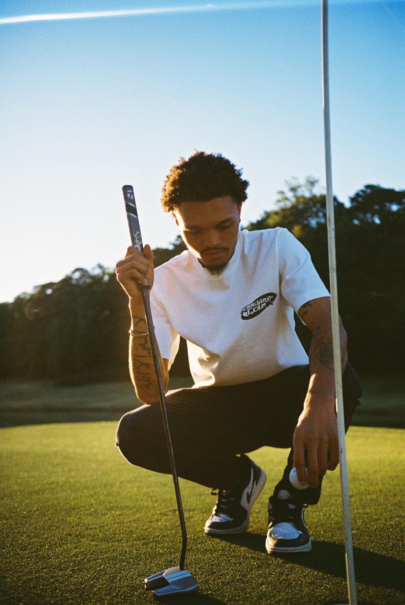Person crouching on grass holding a golf club, with a golf flag nearby and trees in the background.