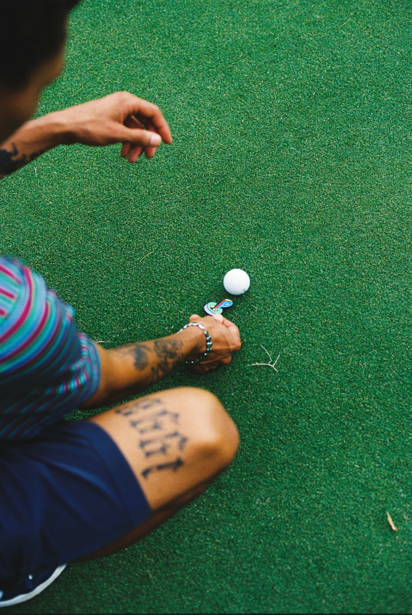 Person kneeling on green surface, preparing to hit a white golf ball with a golf club.