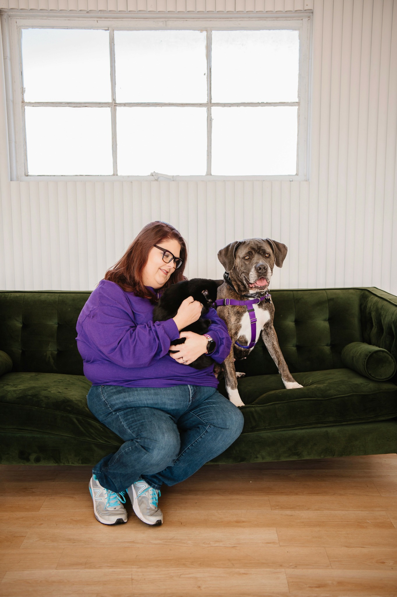 Woman in purple sweater sitting on green sofa with dog wearing purple collar, in room with window and wooden floor.