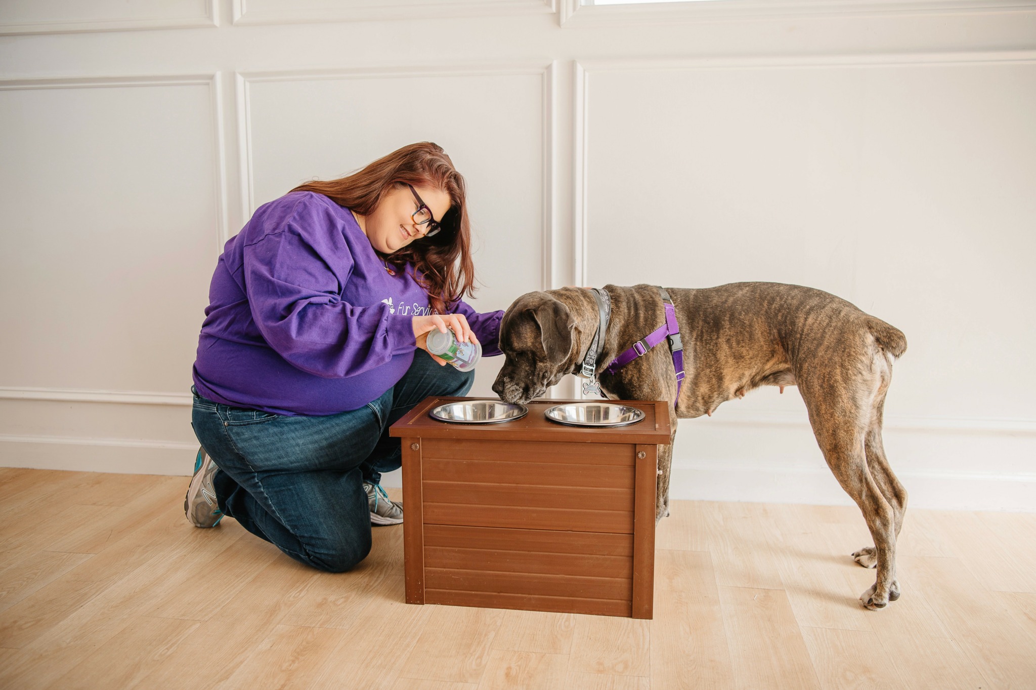 Person kneeling with a dog eating from bowls on a small table in a room with white walls.