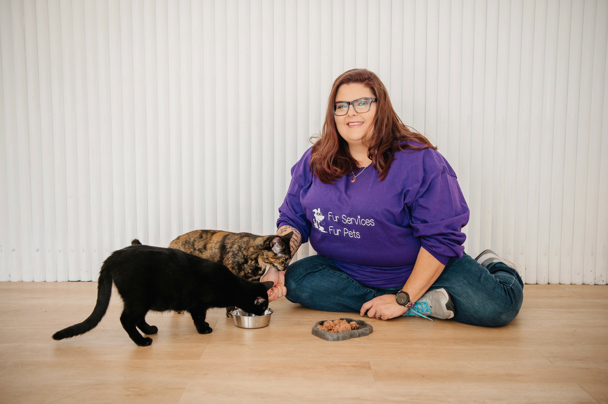 Woman with glasses sitting on floor with two cats and a bowl of food, against a white background.