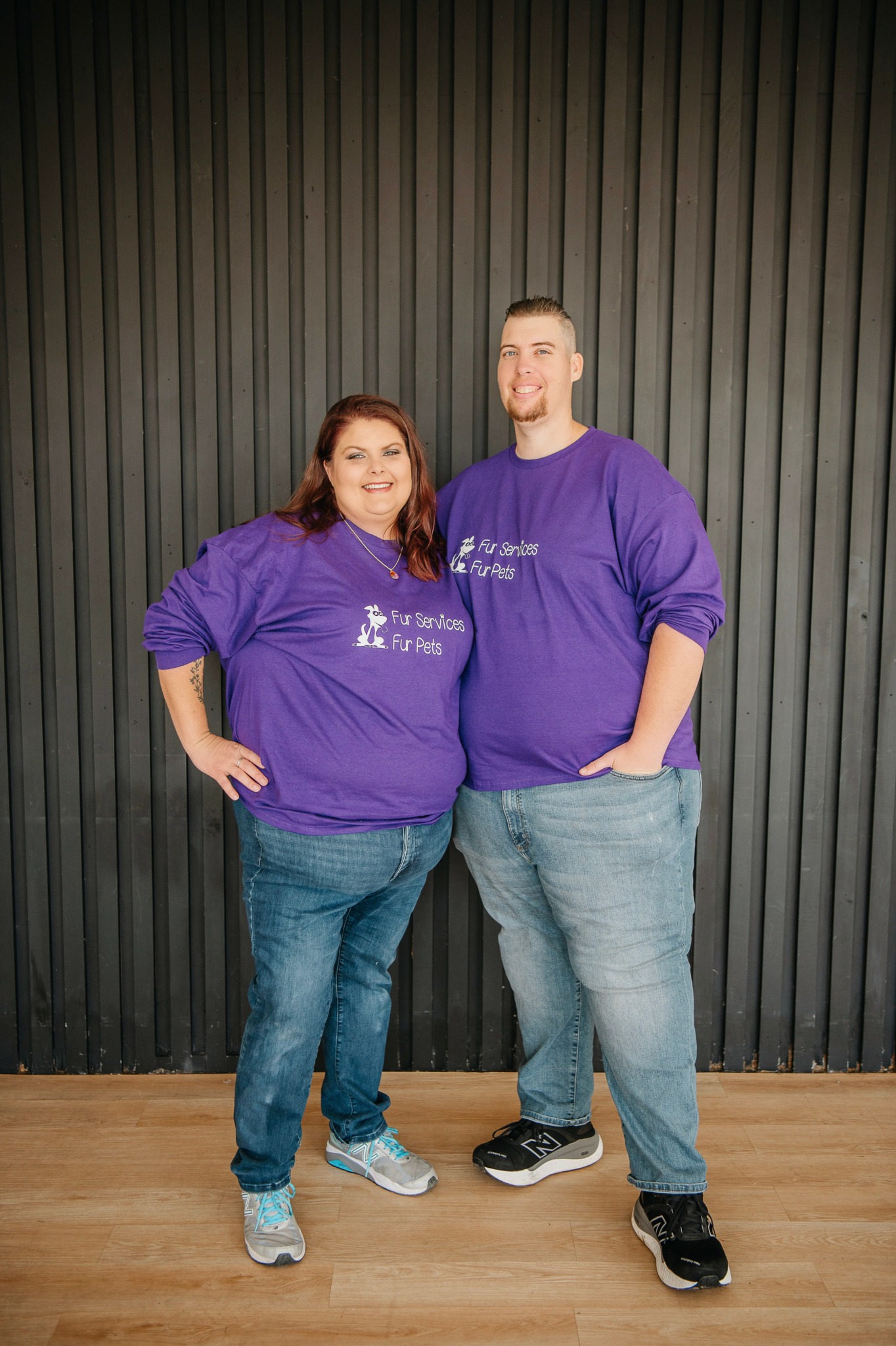 Two people stand side by side in front of a black vertical panel wall, wearing matching purple sweatshirts.
