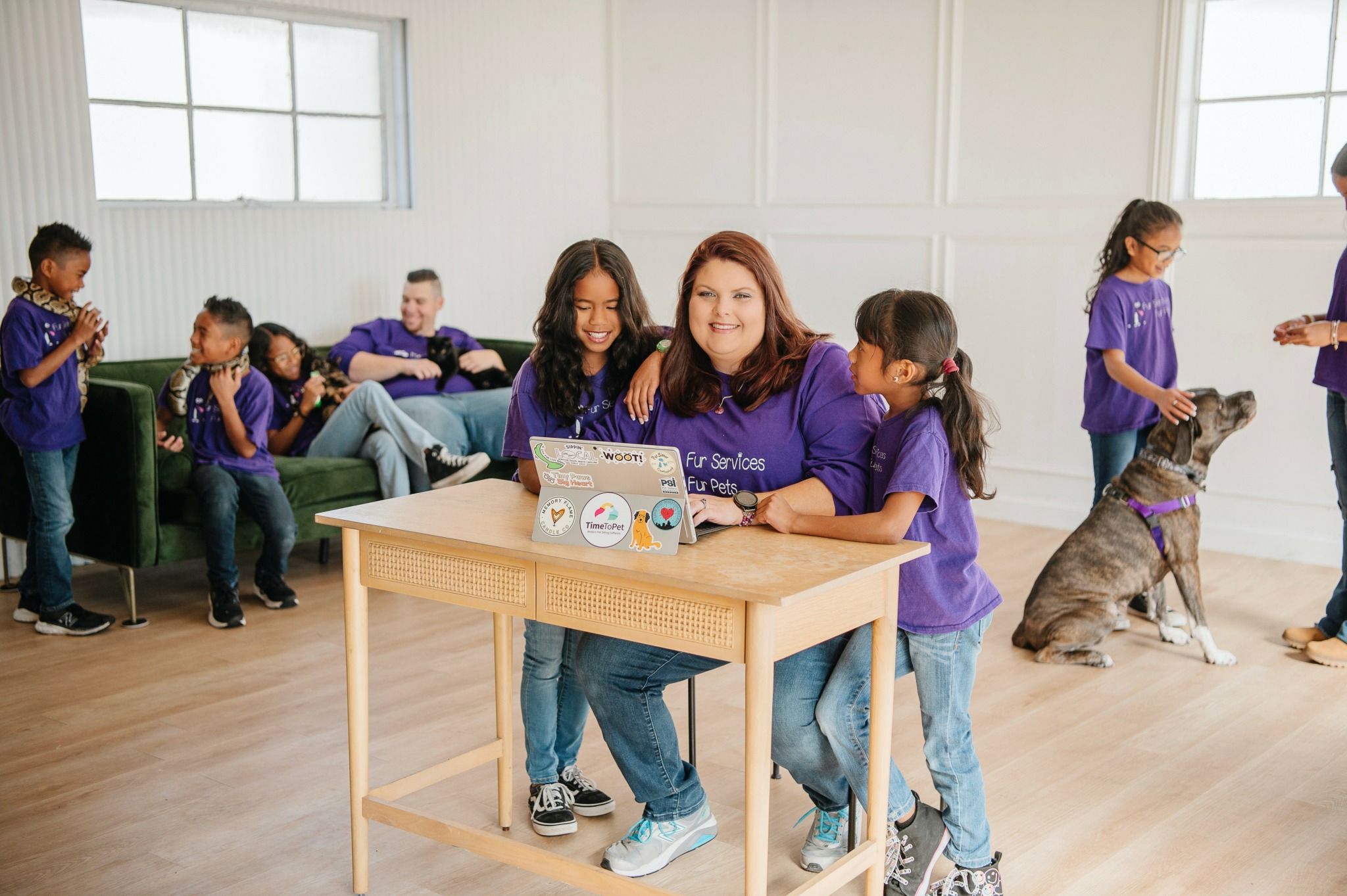 Group of children and a woman in purple shirts in a room with large windows, some sitting, some standing, with a dog.