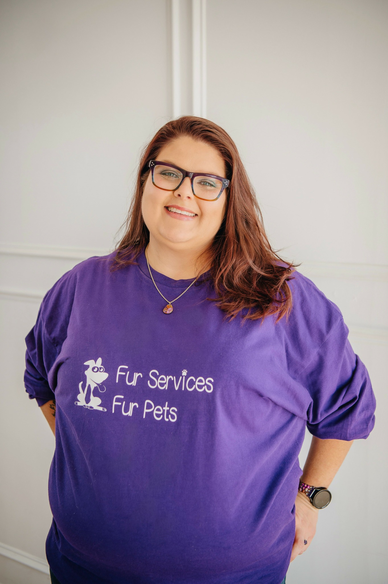 Smiling woman with glasses and long hair wearing a purple Fur Services Fur Pets T-shirt, standing indoors.