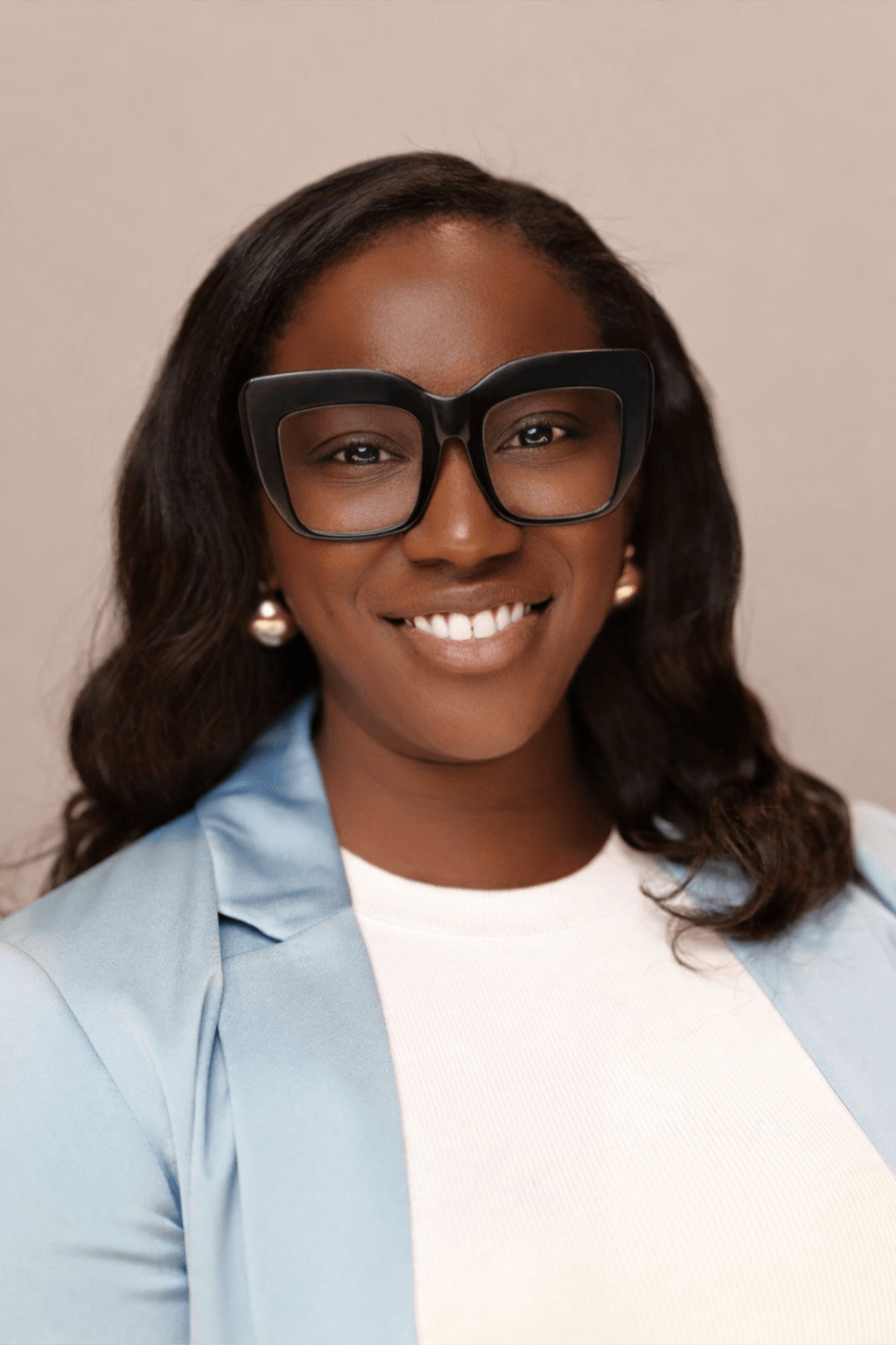 Woman with glasses, earrings, and long wavy hair, smiling, wearing a light blue blazer and white shirt.