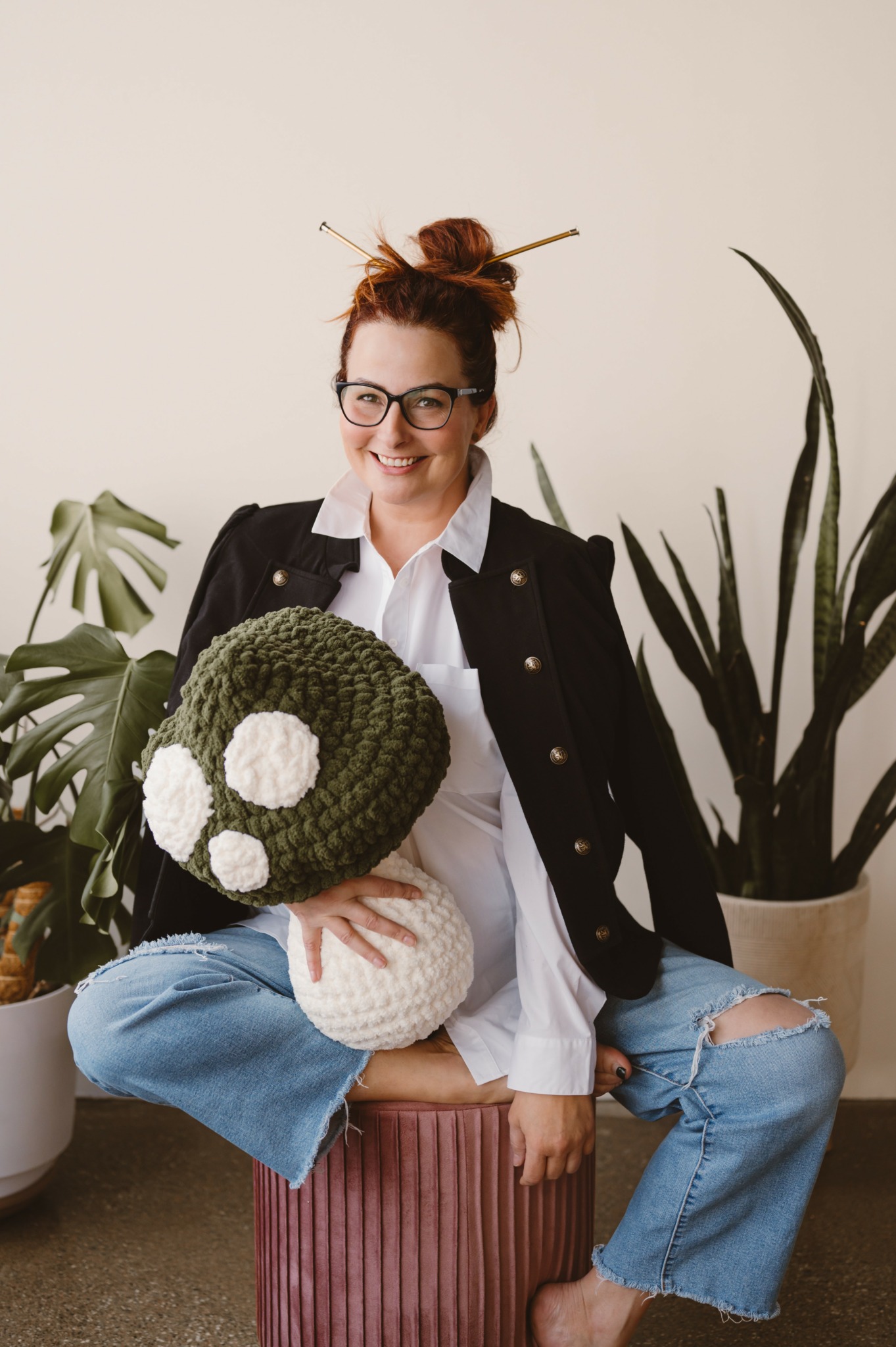 Woman with glasses and red hair in a bun, smiling, holding a knitted green and white plush toy, sitting on a pink stool, surrounded by plants.