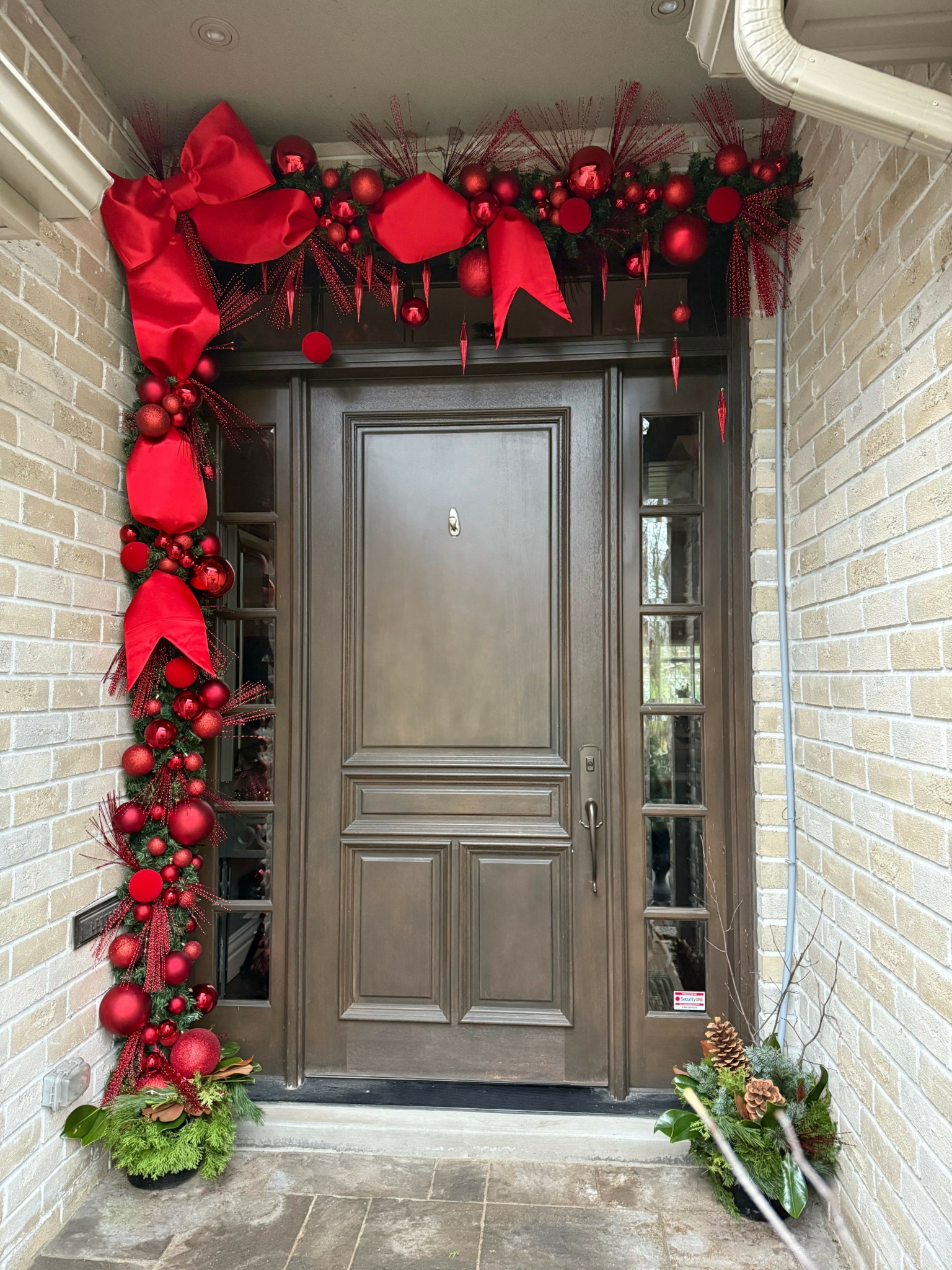 Gray door with Christmas decorations including red ornaments, ribbons, and greenery on a brick wall background.