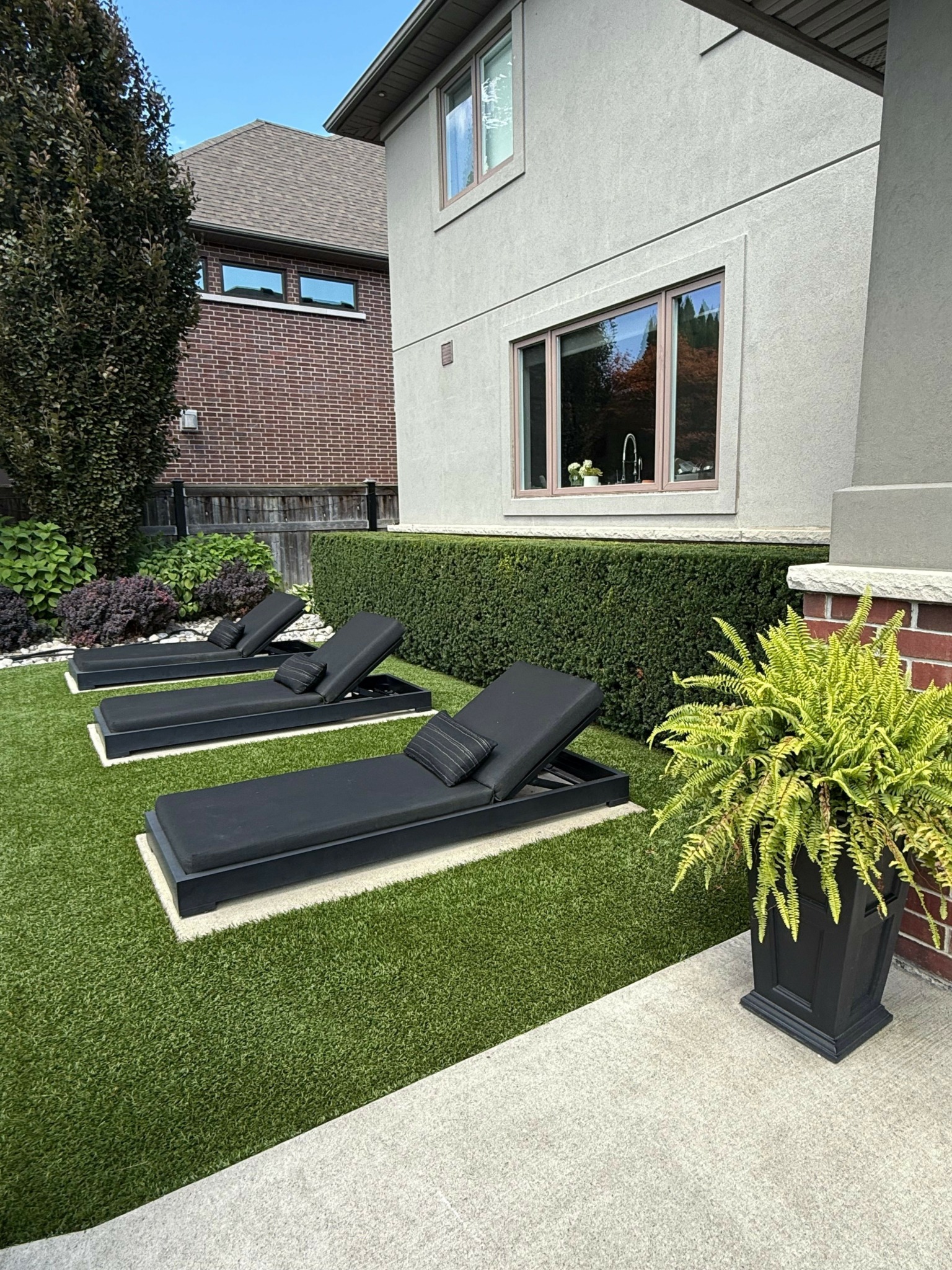 Outdoor patio with black lounge chairs on green artificial grass, potted plant, and building with windows in background.