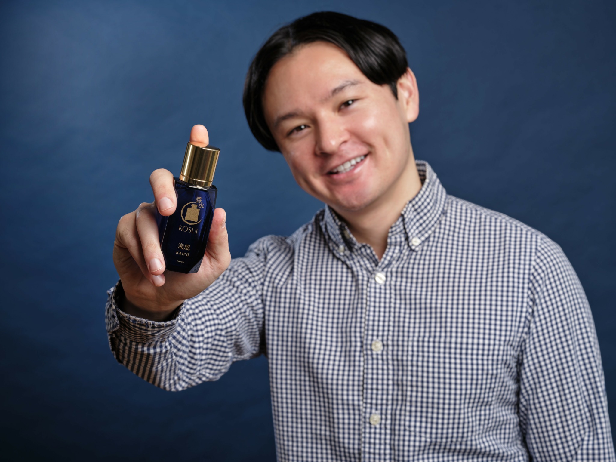 Person smiling holding a small dark bottle with a gold cap against a dark background.