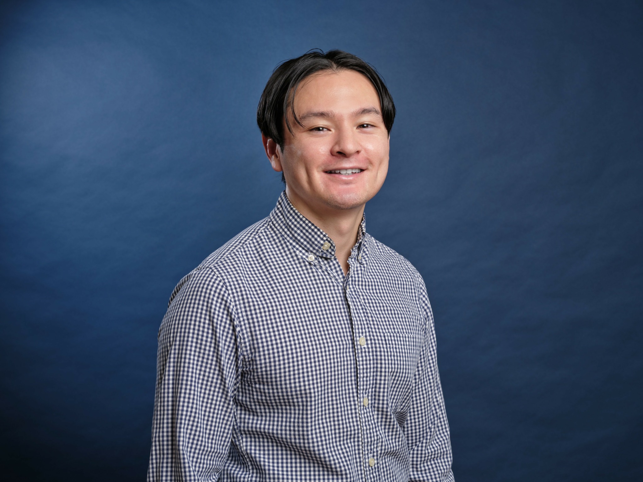 Young man with dark hair smiling, wearing a checkered shirt, standing against a blue background.