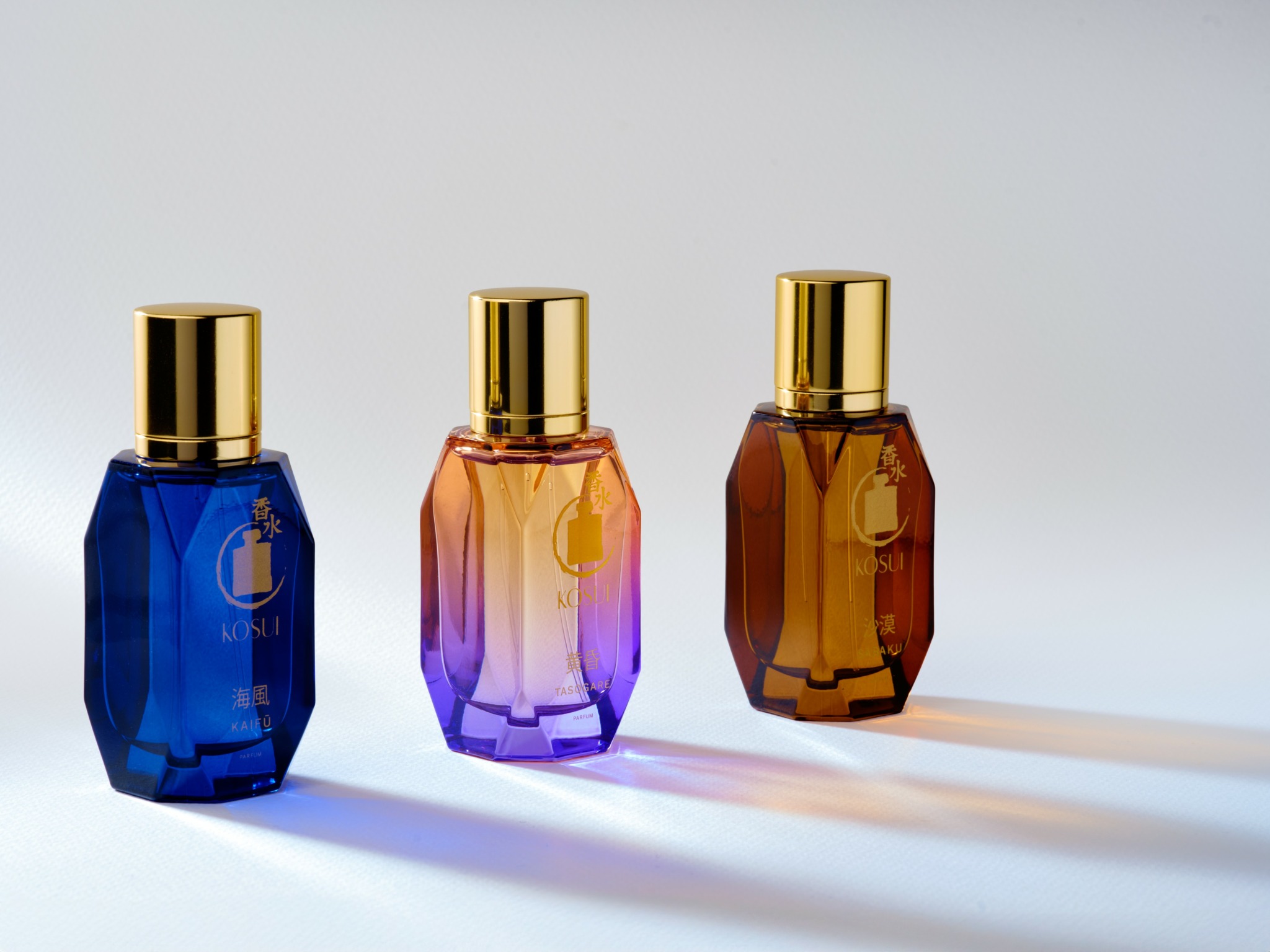 Three colorful perfume bottles with gold caps on a white surface, casting shadows, against a plain background.