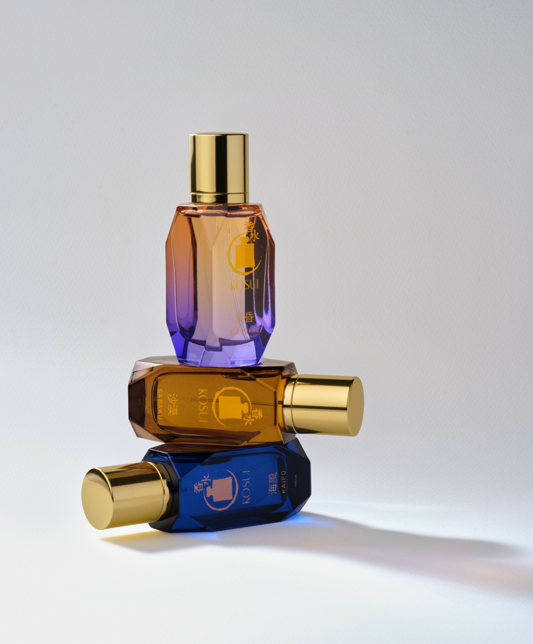 Three colorful perfume bottles with gold caps stacked on a white surface, casting shadows.