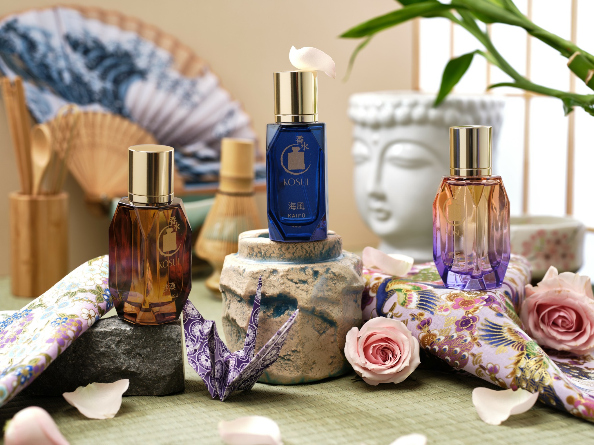 Three perfume bottles on rocks with roses and fabric, indoor setting with plant and decorative items in background.