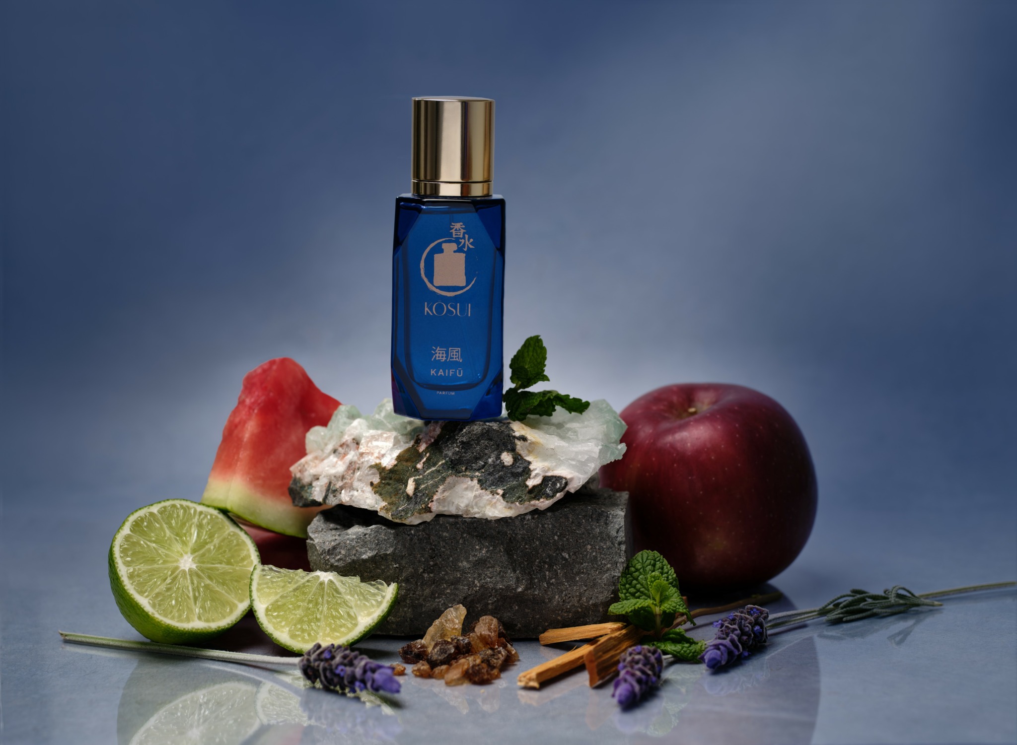 Blue bottle with gold cap on rock, surrounded by watermelon, apple, lime, lavender, cinnamon sticks, and herbs.