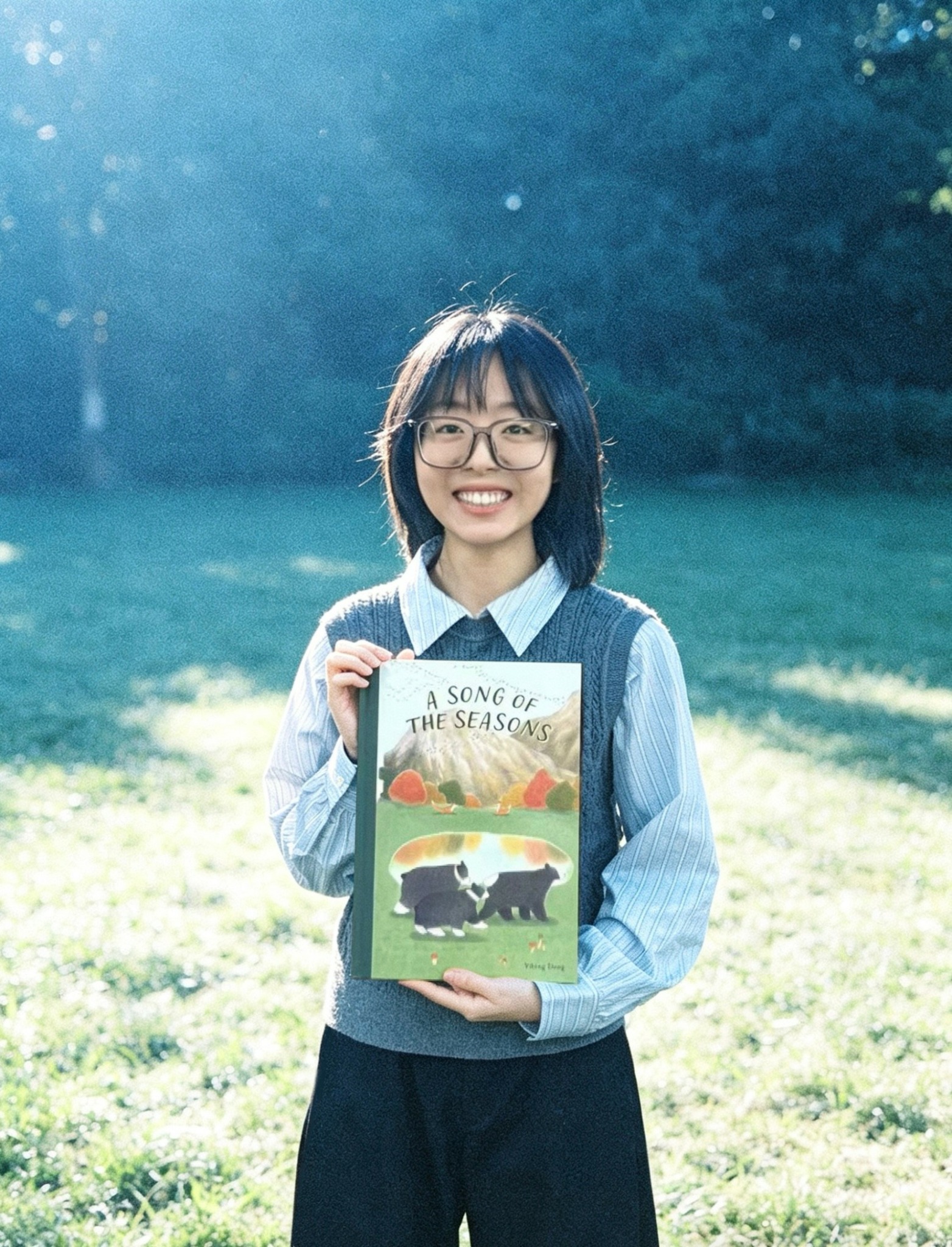 Young woman with glasses holding a book outdoors in a grassy area with trees in background.