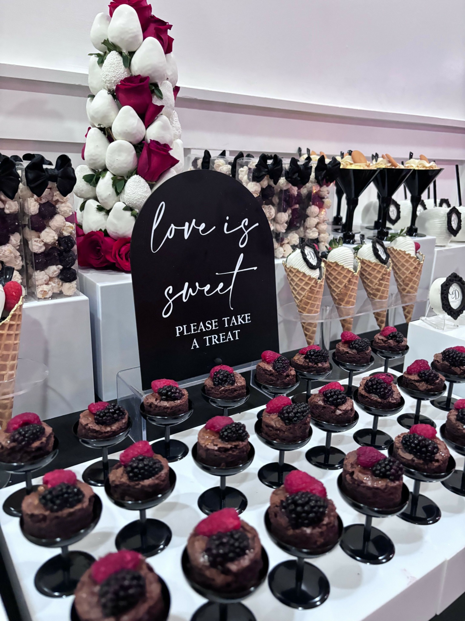 Display of cupcakes with pink and black decorations, a sign with cursive writing, and a floral arrangement in a bakery.