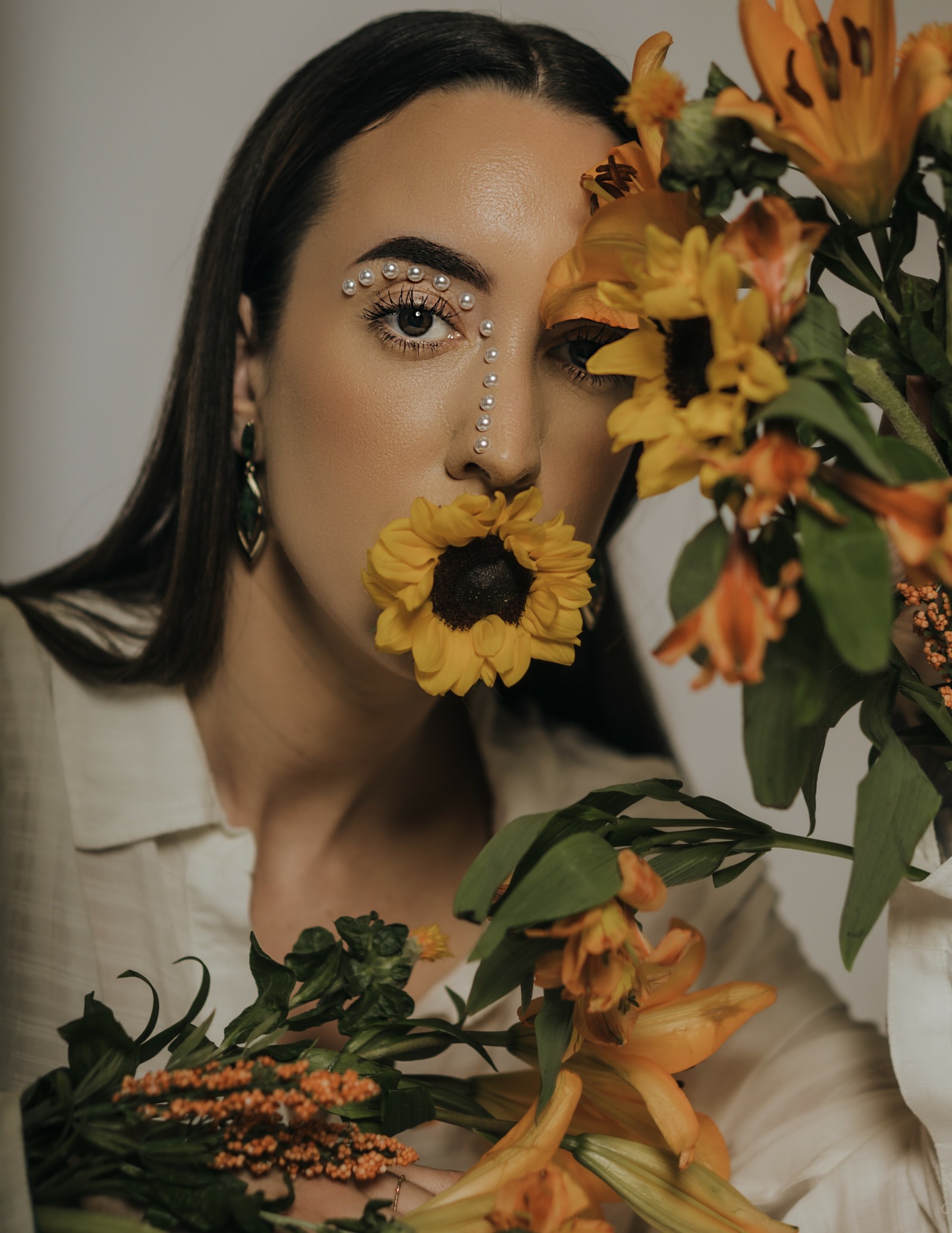 Woman with dark hair and decorative face jewelry partially hidden behind a bouquet of yellow and orange flowers.