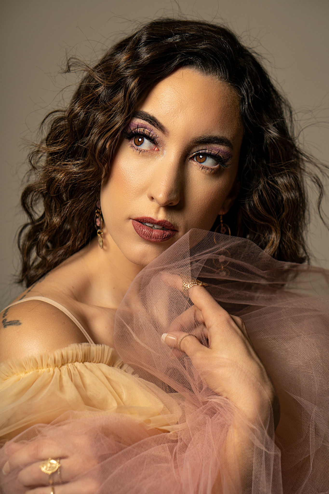 Woman with curly hair and makeup, holding sheer fabric near face, wearing a yellow off-shoulder top.