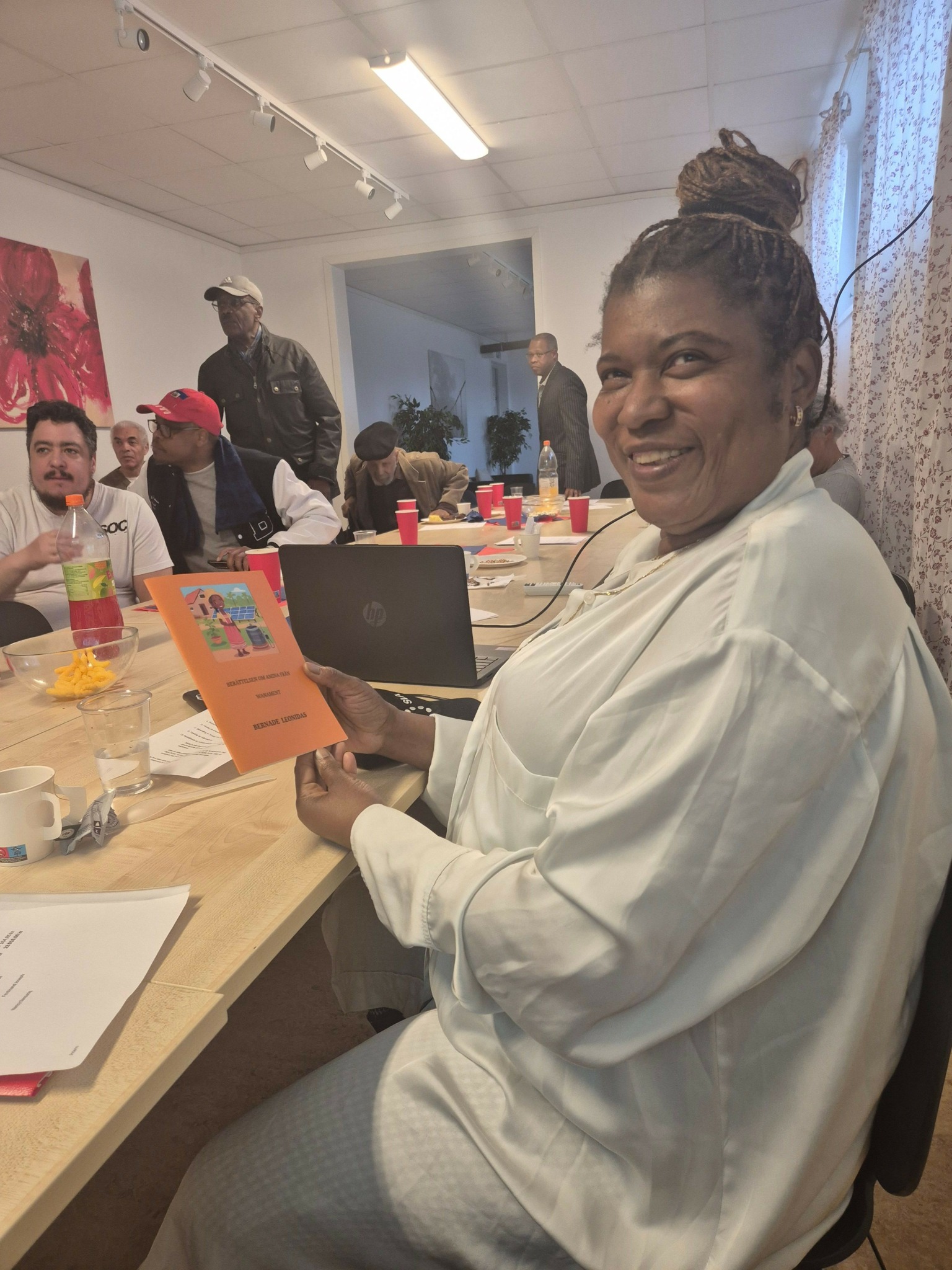 Woman in white coat smiling at camera, standing at table with colorful items and papers, in a room with white walls.