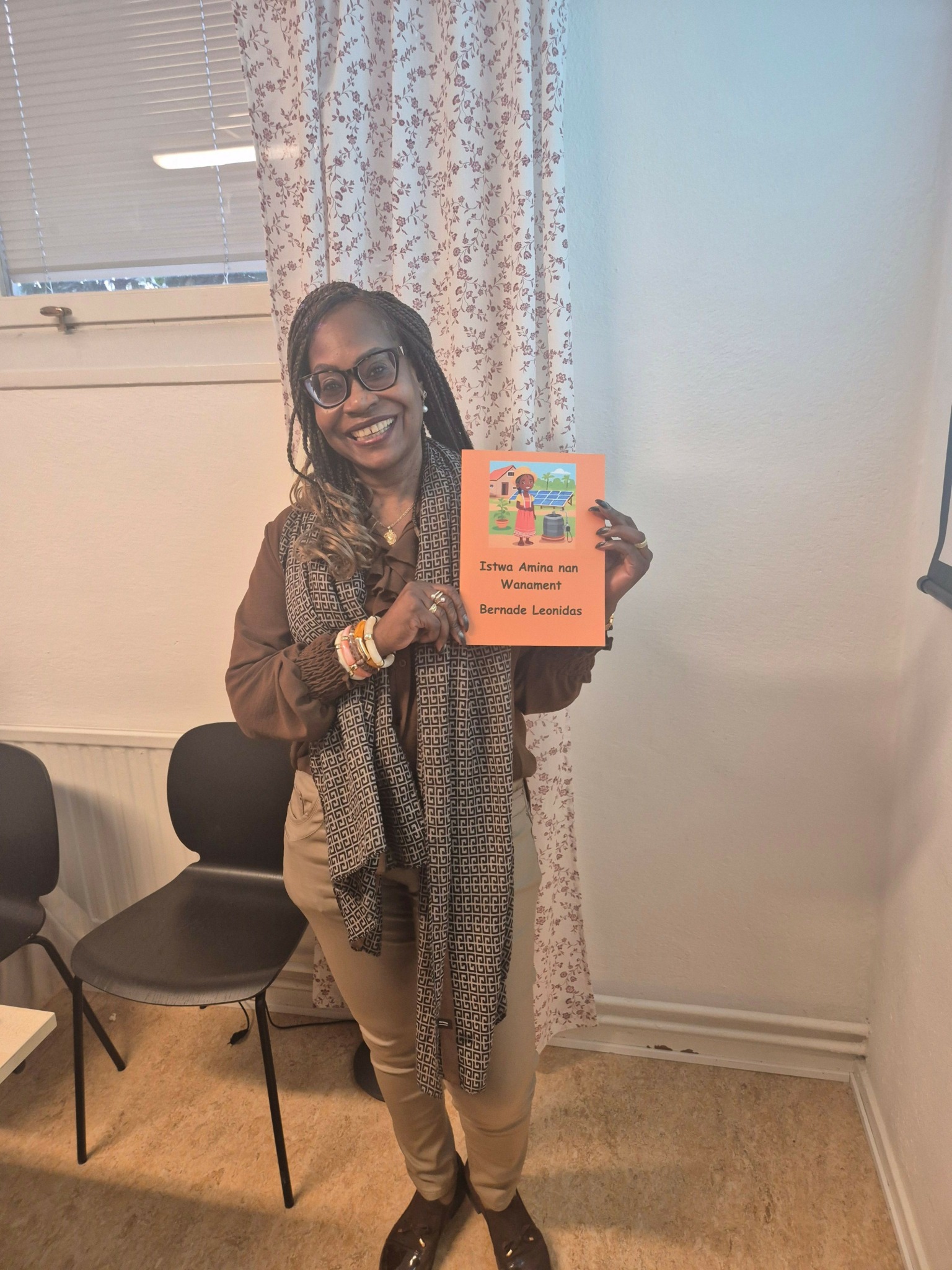 Woman with glasses holding a colorful card, standing in a room with curtains and a chair.