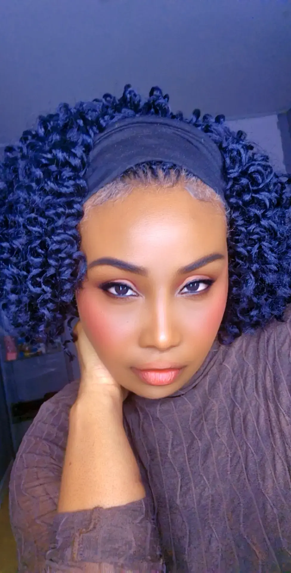 Close-up of a woman with curly hair wearing a headband, resting her face on her hand, looking at the camera.