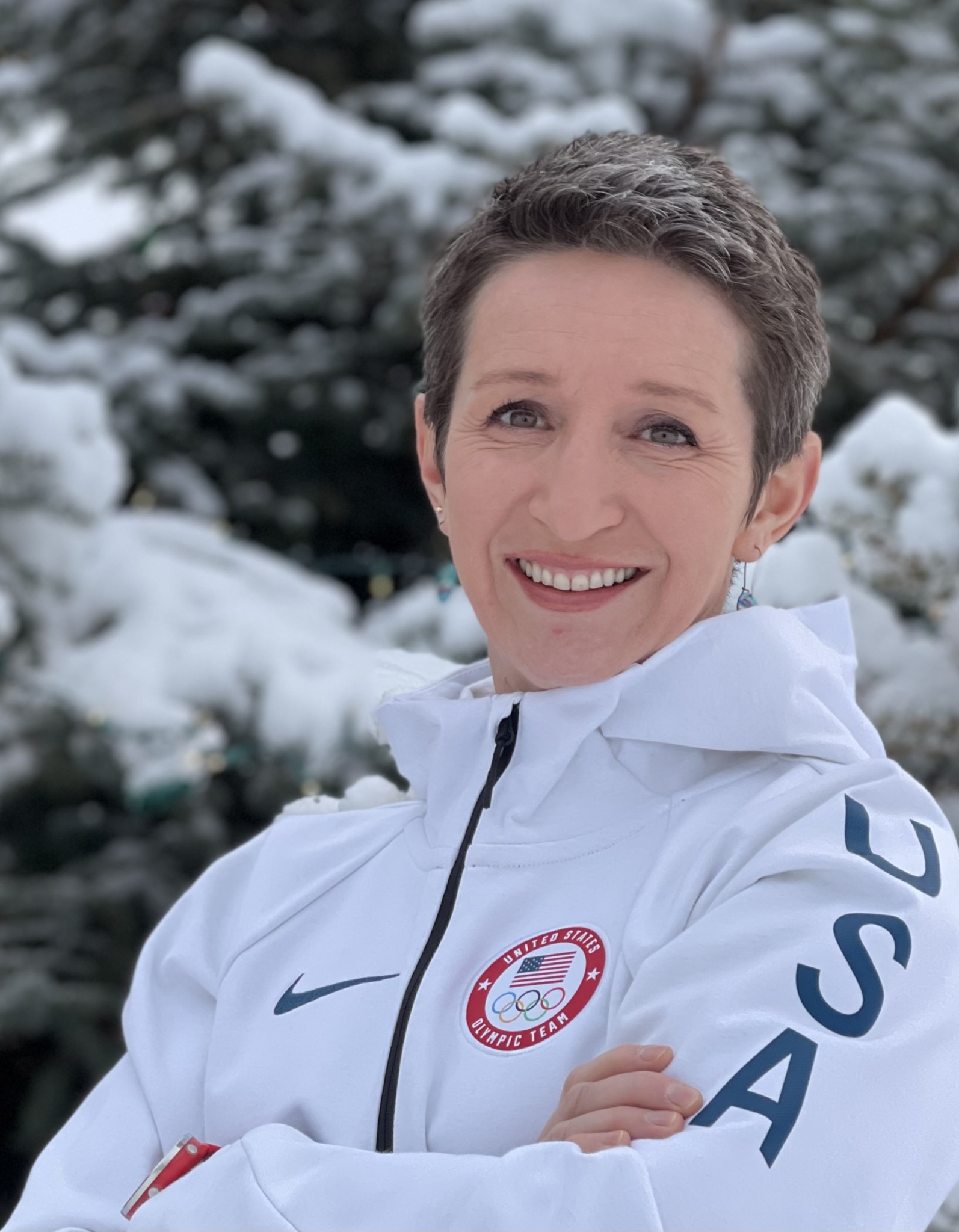 Woman smiling outdoors in winter, wearing a white jacket with USA and Olympic logos, snow-covered trees in background.