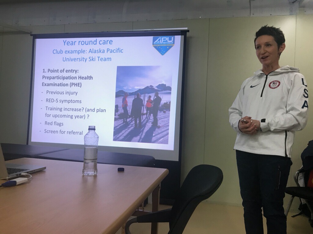 Person standing next to a presentation slide about year-round care and ski team, with a table and chair in foreground.