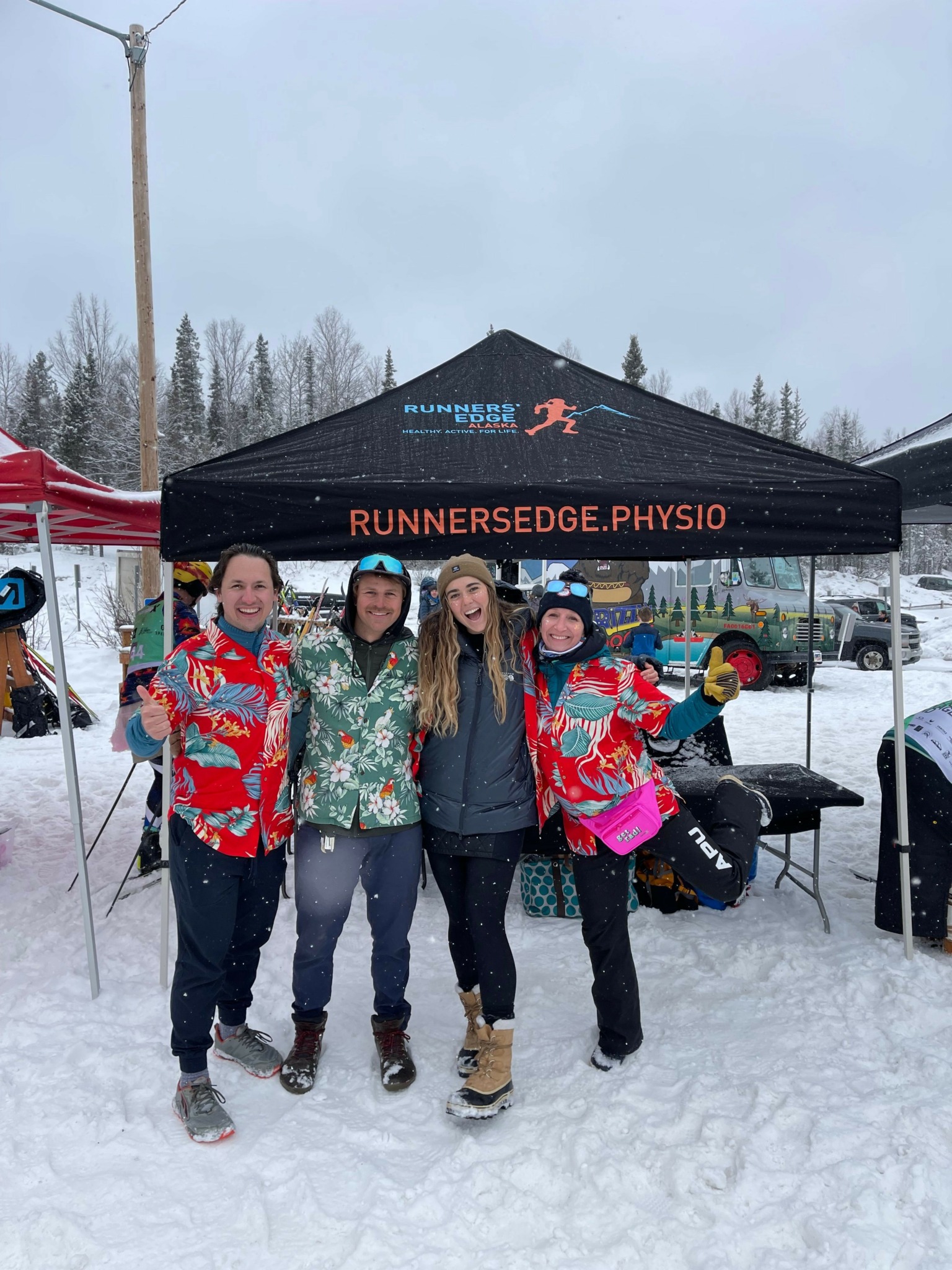 Four people standing in snow near a sign that reads 'RUNNERSBRIDGE PHYSIO'. They wear winter clothing and masks.