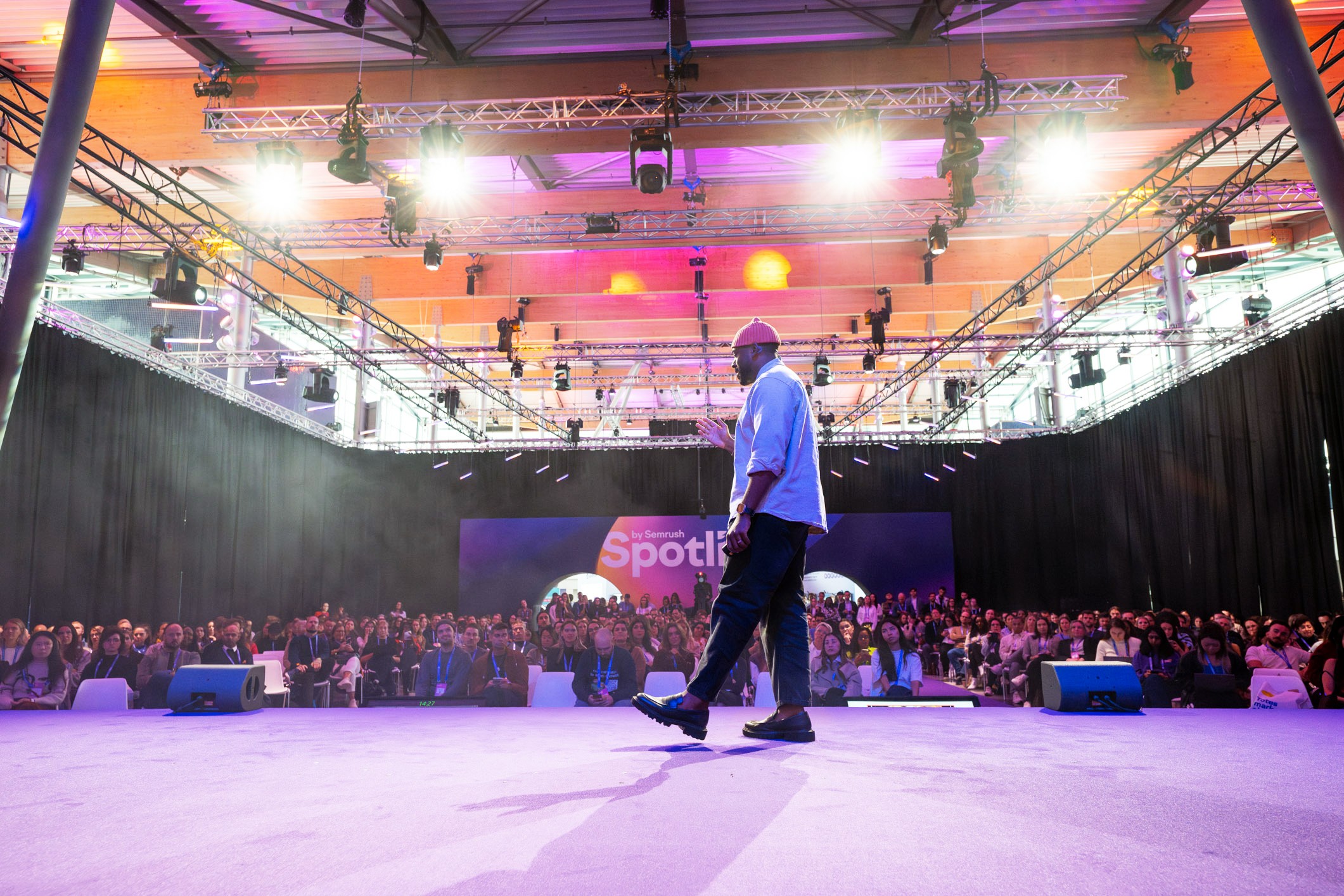 Person walking on stage with a large audience and colorful lighting overhead.
