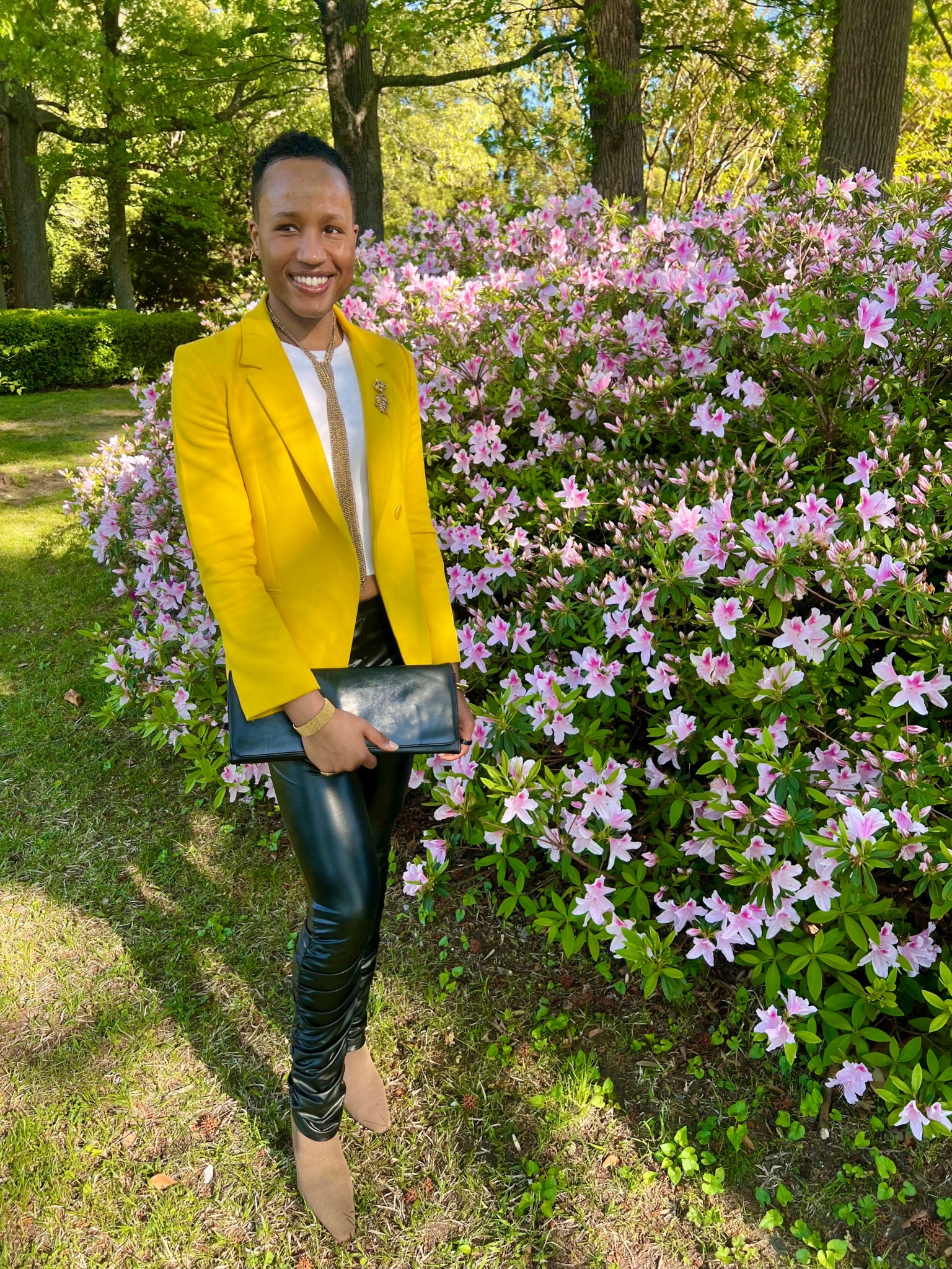 Woman in yellow blazer and black pants standing next to pink flowering bush in park.