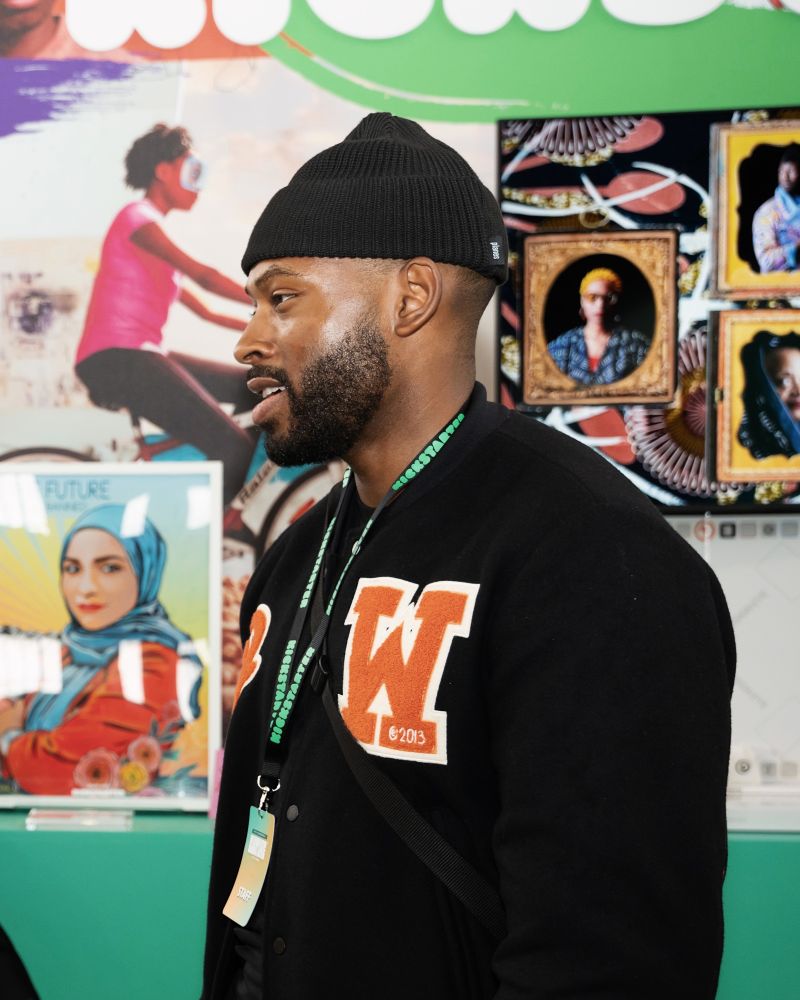 Man wearing a black beanie and black jacket with a graphic patch, looking to the left, in an indoor setting with colorful displays.