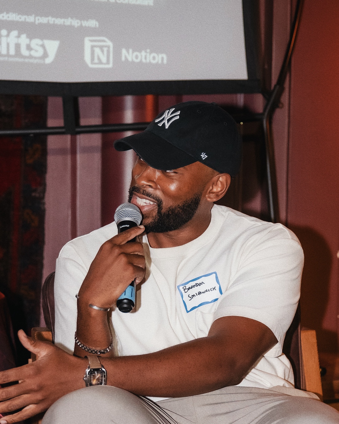 Man with a beard wearing a black cap and white t-shirt, smiling and speaking into a microphone.
