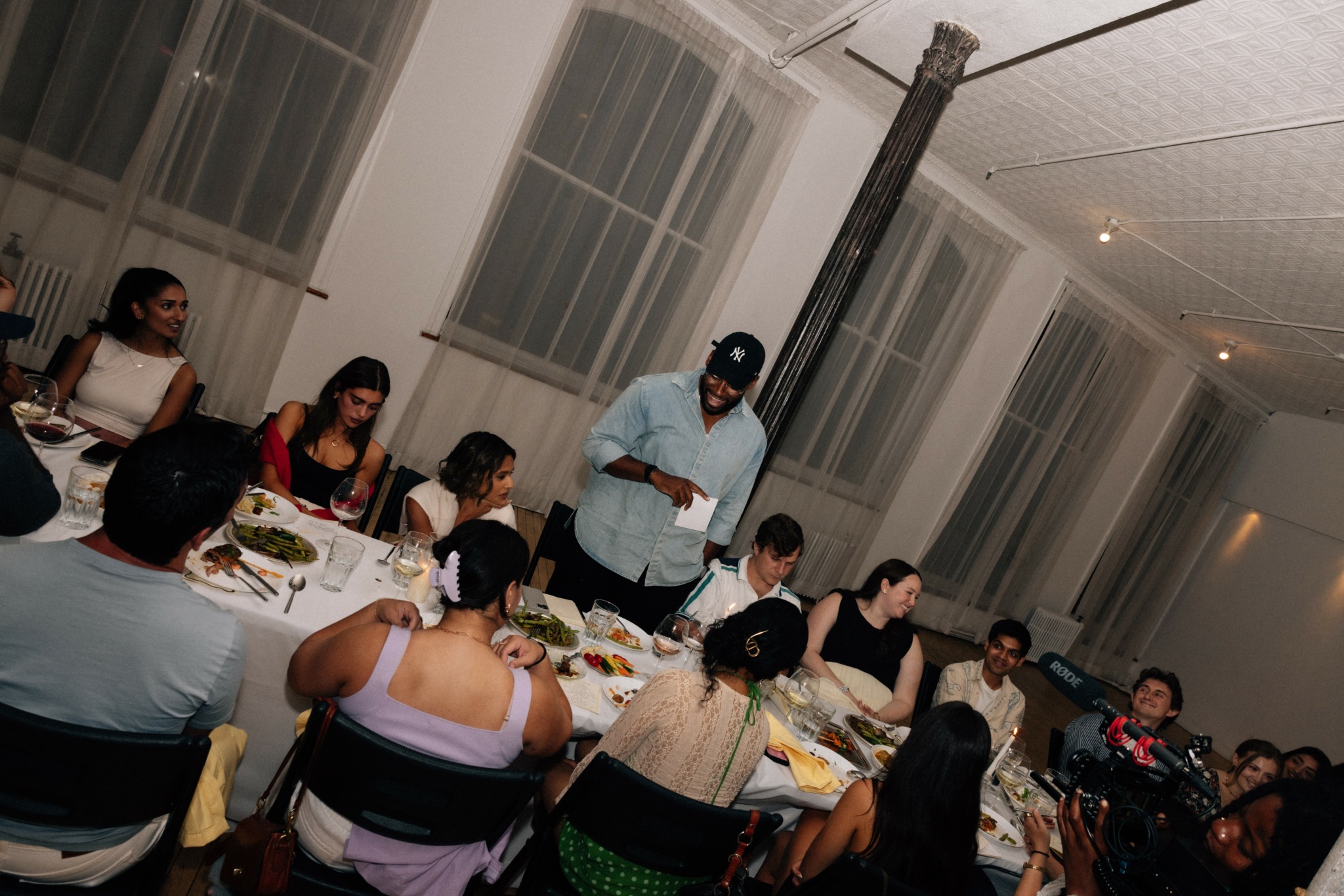 People seated at a long table with food, in a room with large windows and white curtains.