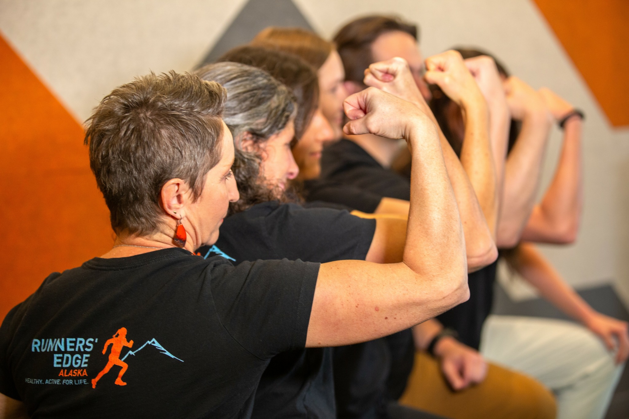 Group of people in a line, flexing their arms and showing muscles, indoors with a neutral background.