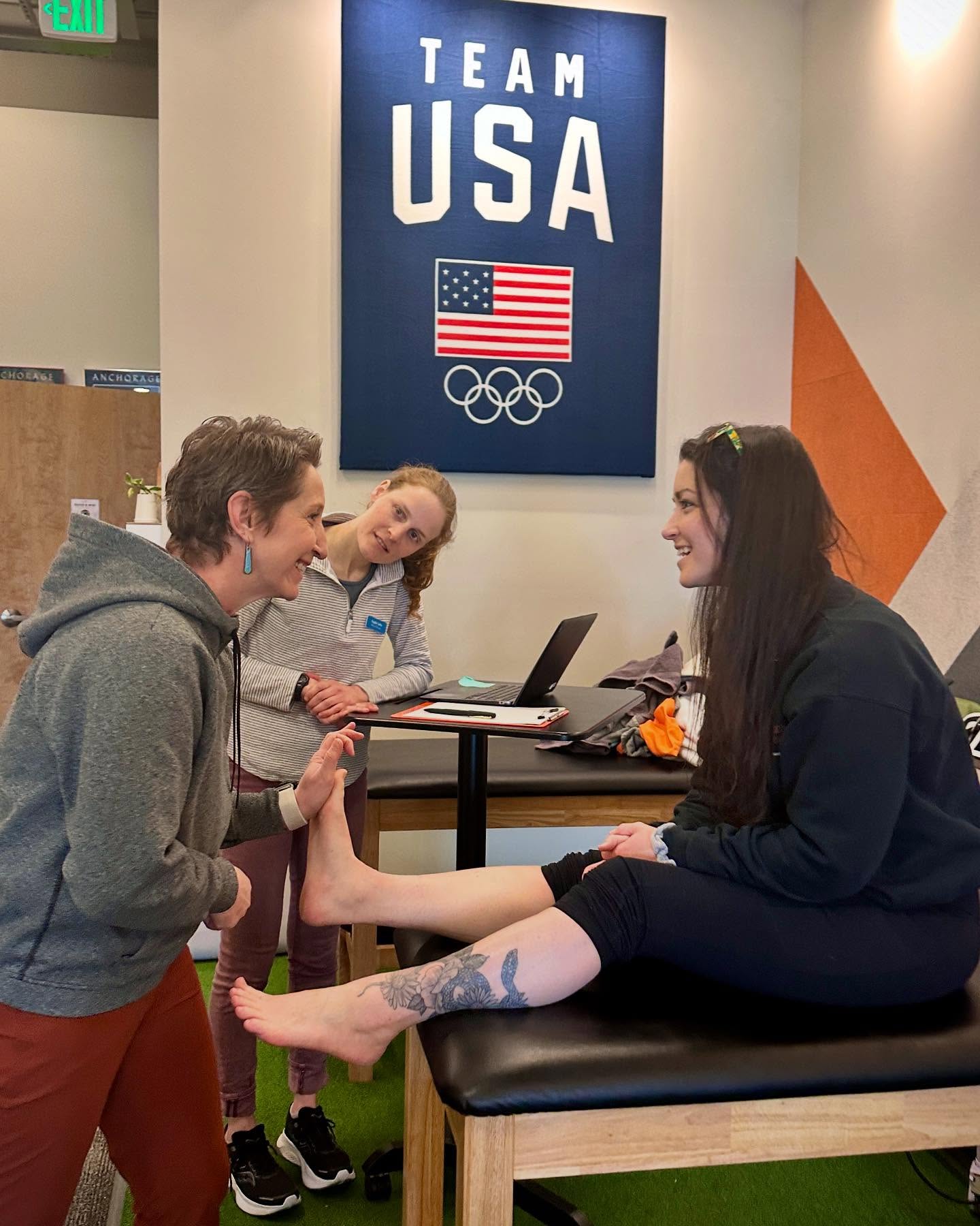 Two women and a girl in a room with a 'Team USA' Olympic sign on the wall. One woman is sitting on a table, the other standing, holding the girl's hand. The girl is standing between them, looking at the woman sitting.