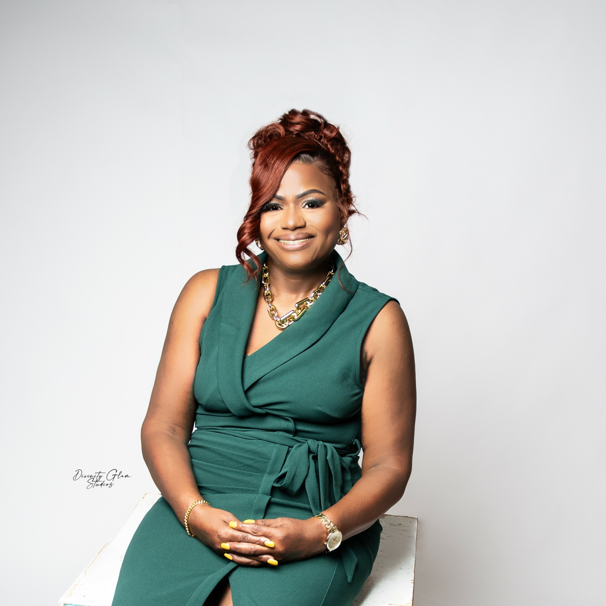 Woman with red hair in a green sleeveless dress, smiling, sitting on a white surface, against a plain white background.