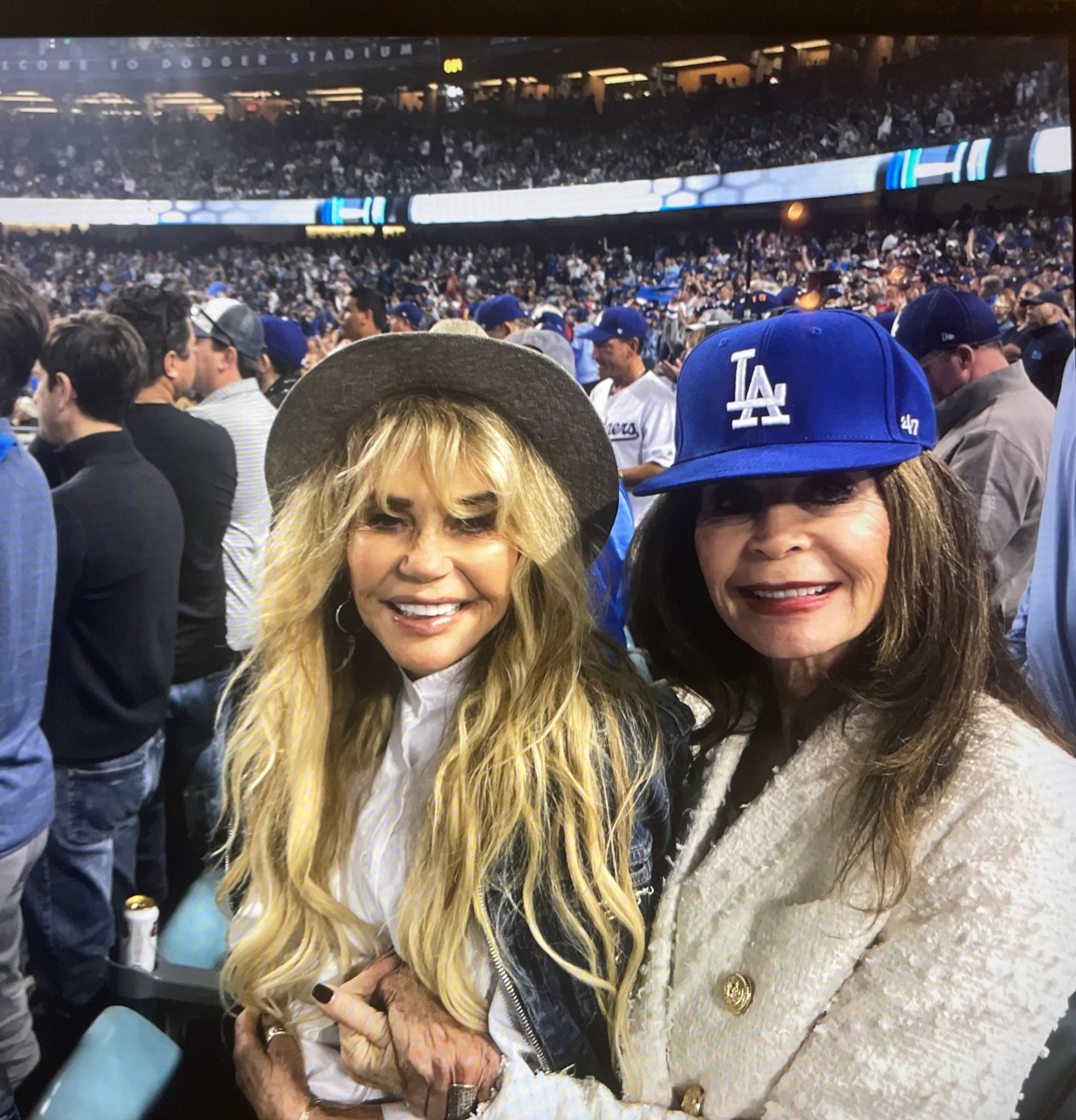 Two women smiling at a stadium, one with long blonde hair and the other with long dark hair, both wearing hats.