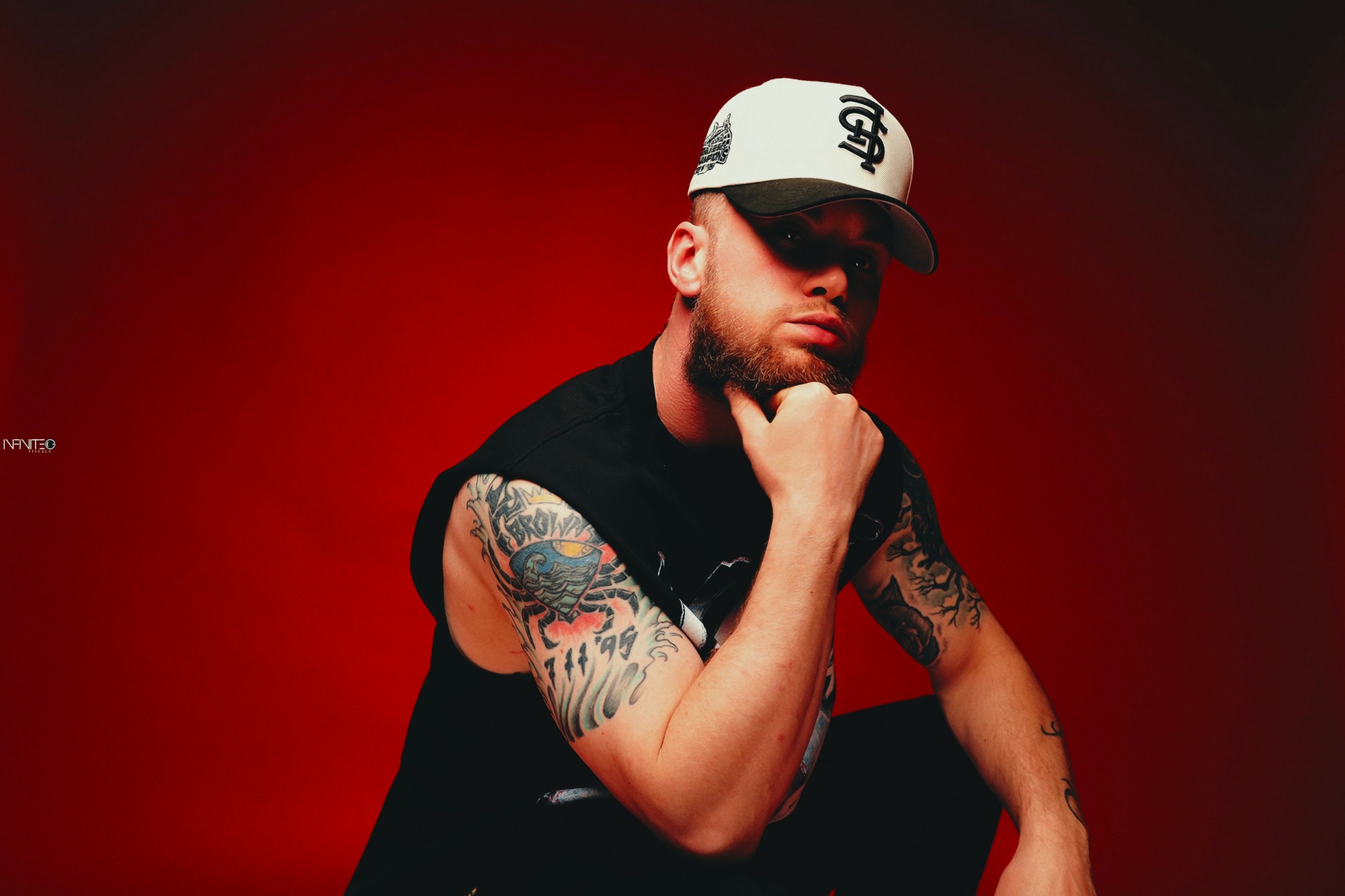 Man with tattoos wearing a cap and black shirt, resting chin on hand, against red background.