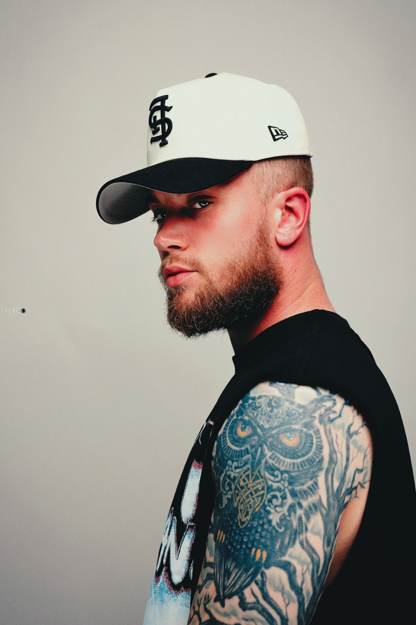 Young man with a beard wearing a white cap and sleeveless shirt, showing a tattooed arm, against a plain background.