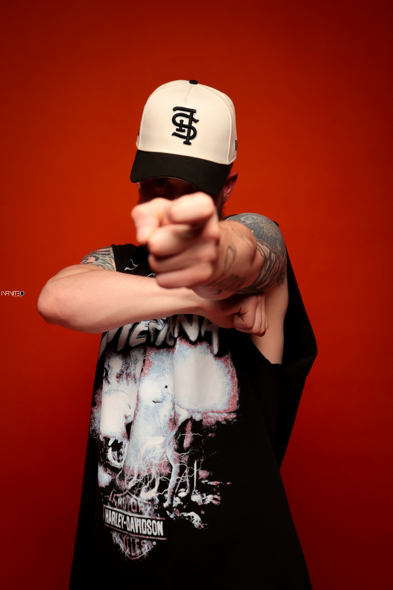 Person wearing a white cap with a logo, pointing directly at the camera, with tattoos on arms, against an orange background.